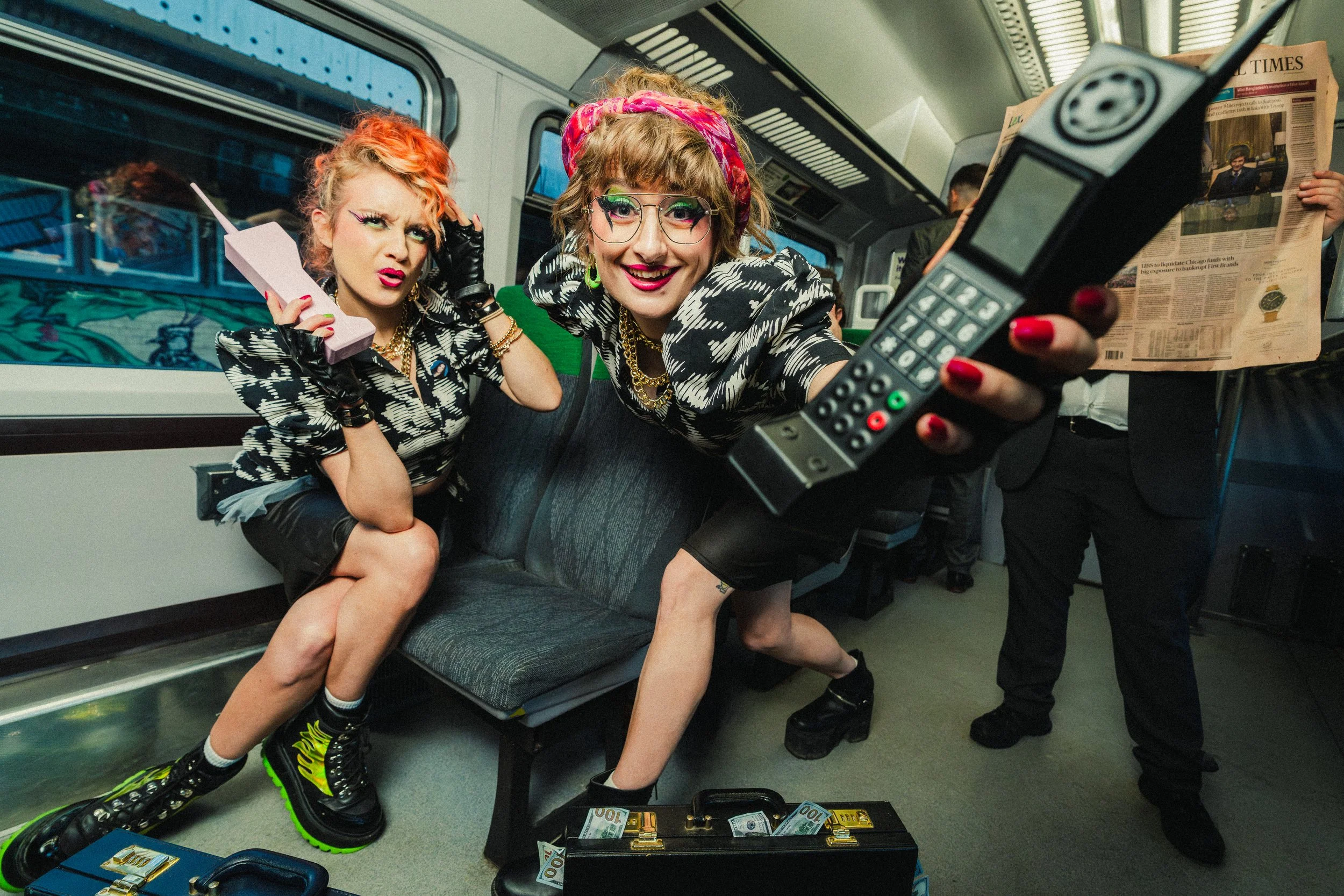 Two women dressed in edgy, vintage-style clothing with colorful hair and makeup, sitting and standing on a bus, posing with a large remote control and a pink flip phone, surrounded by bags with cash and a man in the background reading a newspaper.