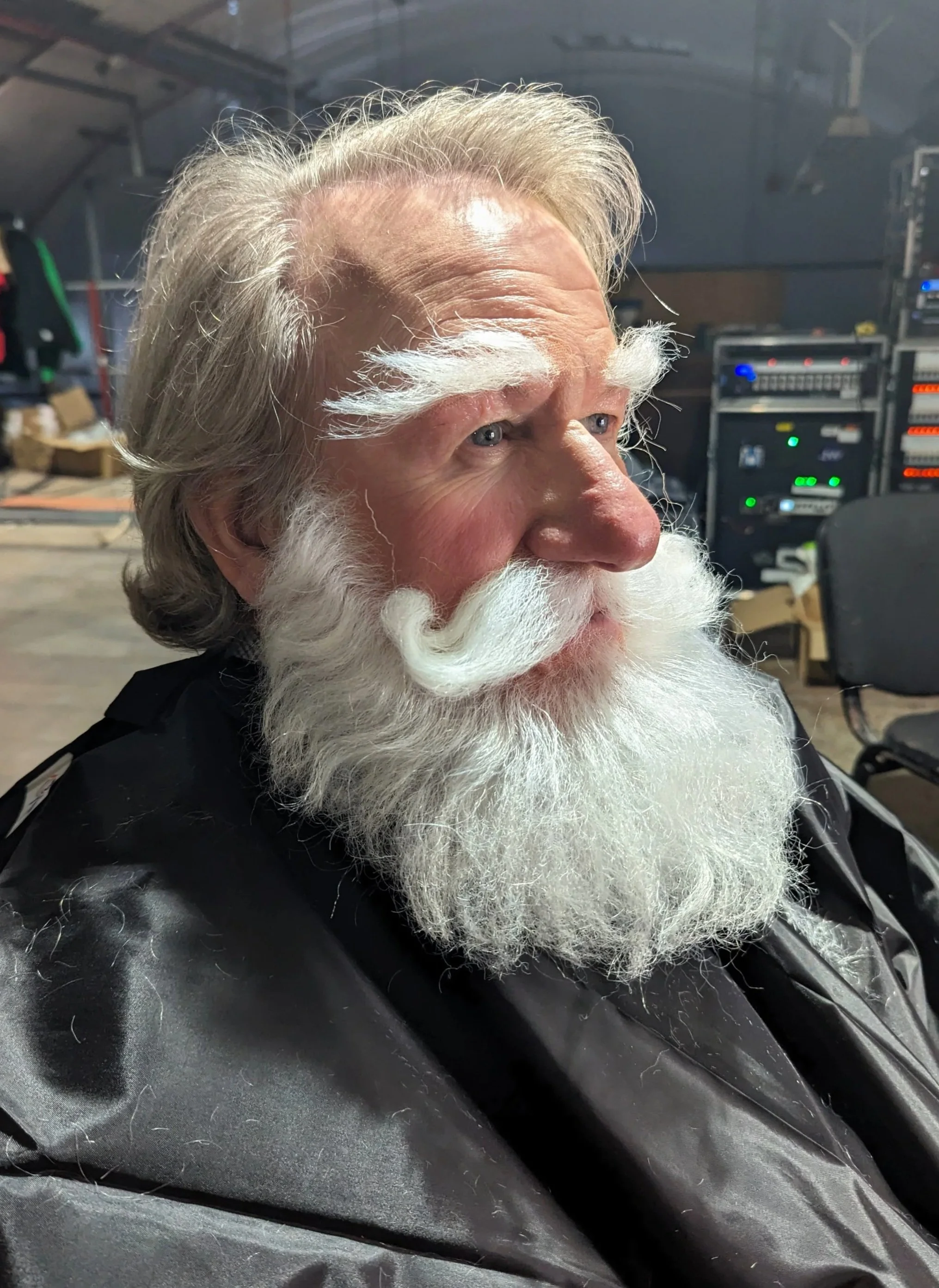 A close-up side profile of an elderly man with a white beard and white, bushy mustache, sitting in a room with electronic equipment in the background.