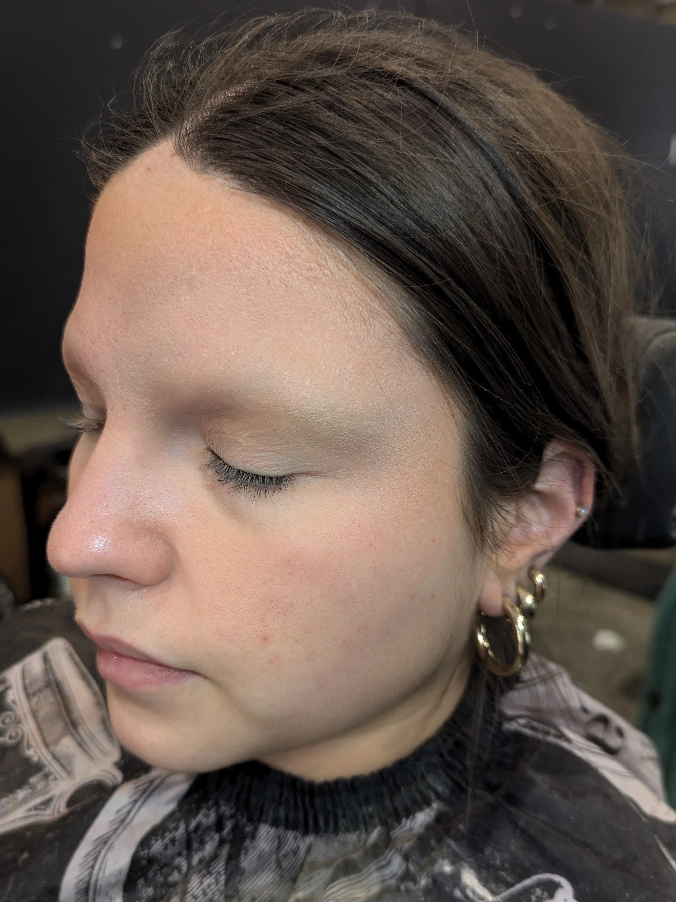Close-up of a woman with a smooth, makeup-free face, side profile, with dark brown hair and multiple gold earrings, sitting in a hair salon.