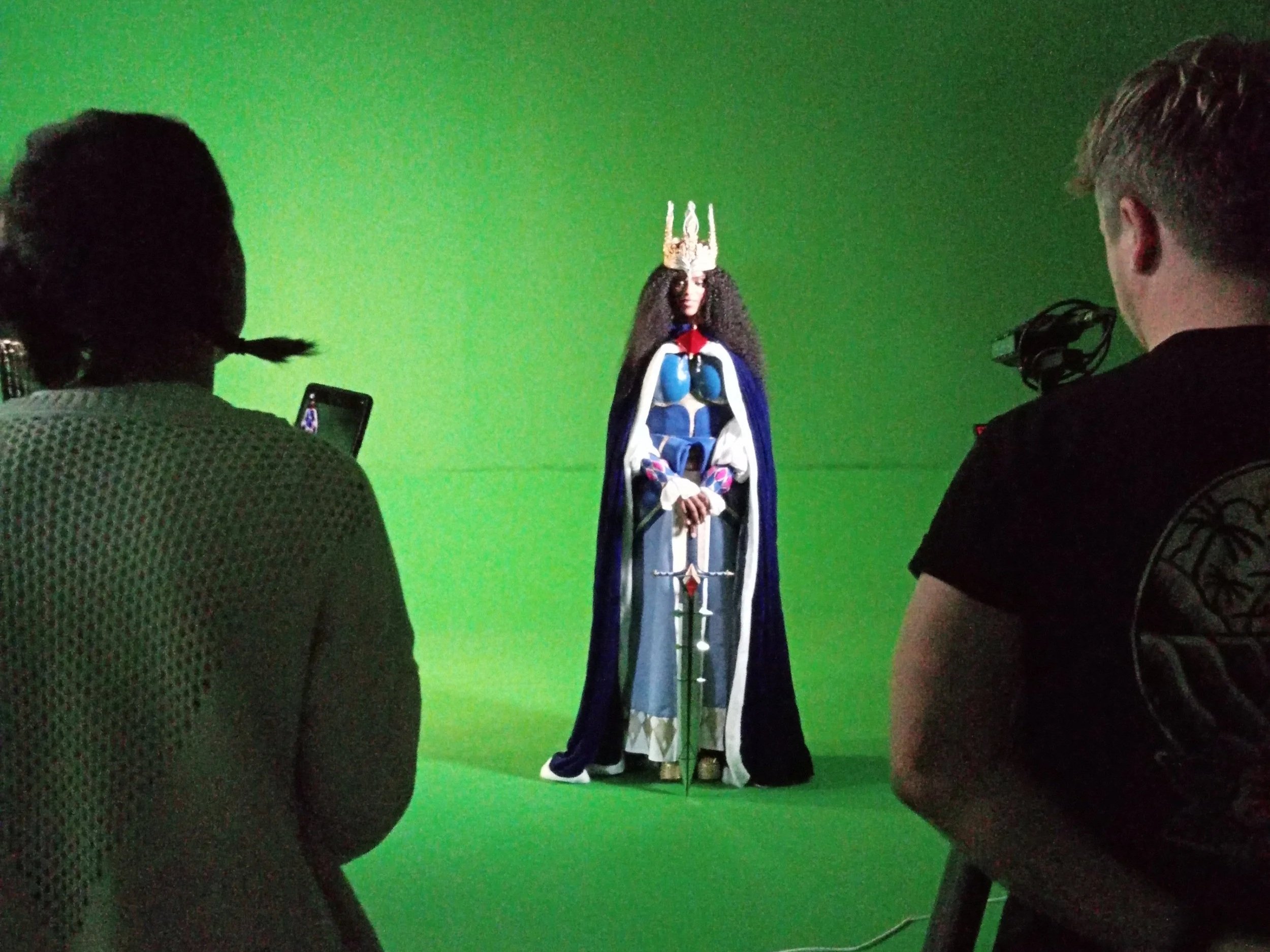 Behind-the-scenes of a film or photoshoot with green screen, featuring a woman dressed as a fantasy character with a crown, cape, and sword.