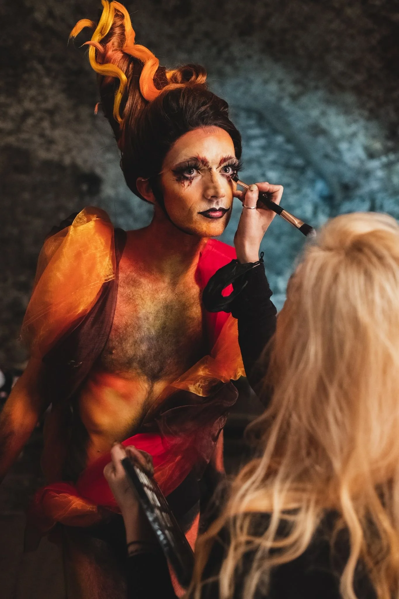 A person with elaborate, multicolored hair and artistic makeup is applying makeup to another person's face. The person being painted has a colorful, theatrical costume.