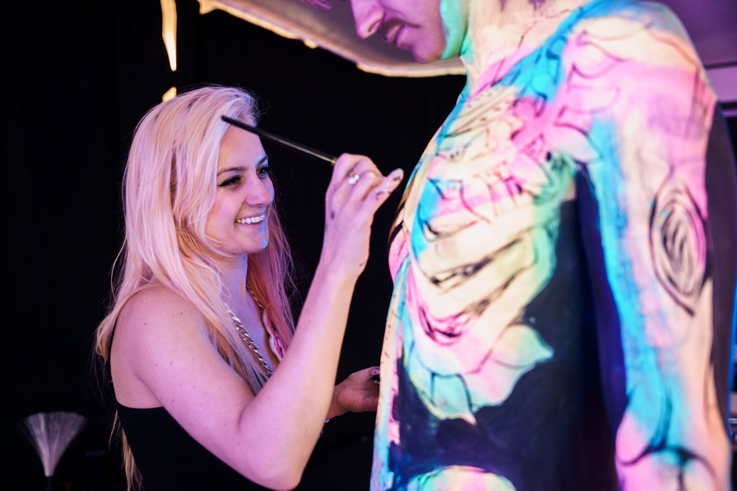 A woman with long pink hair smiling as she paints a colorful, abstract tattoo design on a person's arm with a brush. The person being painted is wearing a black shirt and the background is dark.
