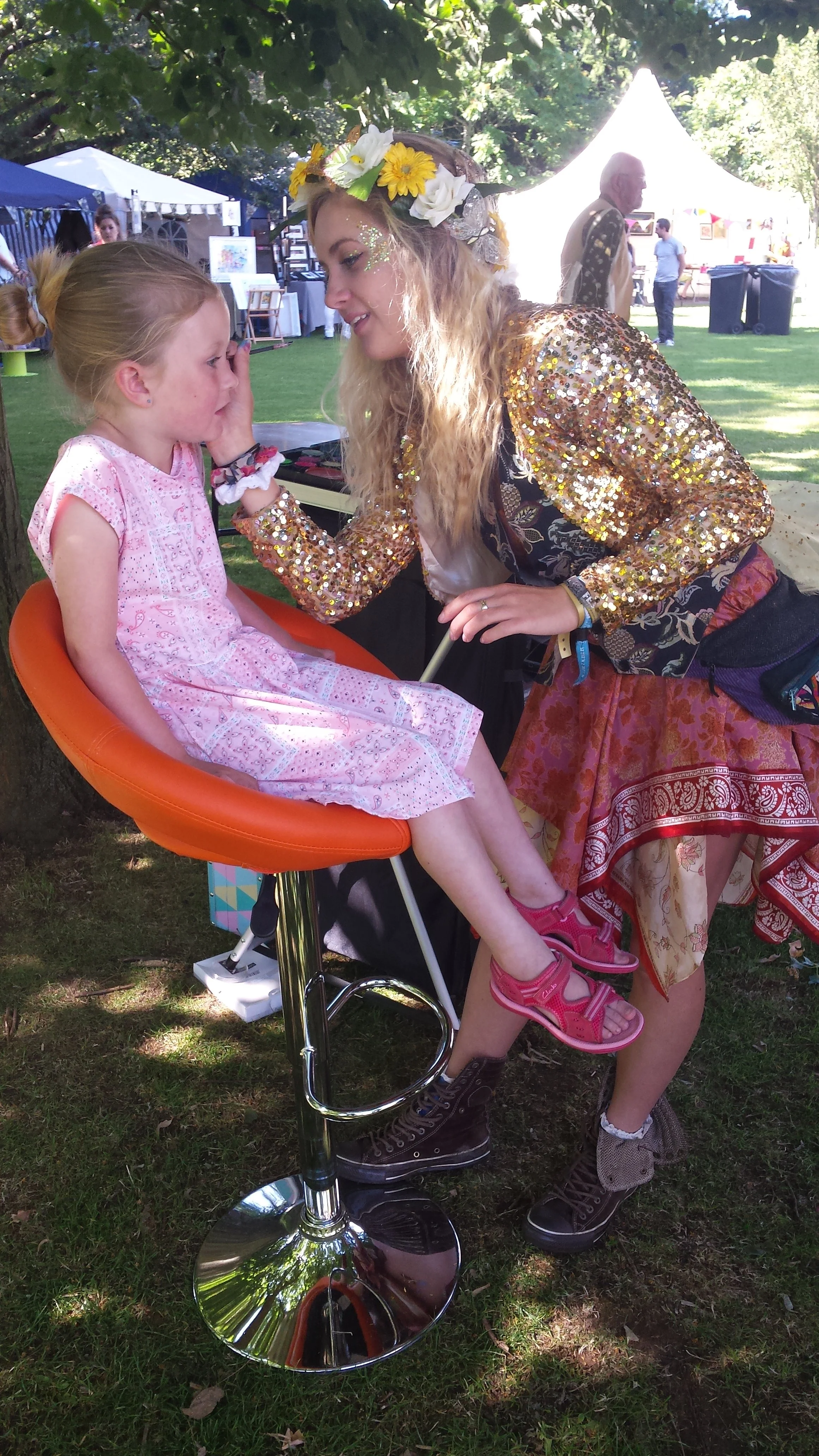 A young girl with a pink dress is sitting on an orange stool, having her face painted by a woman wearing a flower crown, gold sequined jacket, and colorful skirt at an outdoor event with tents and trees.
