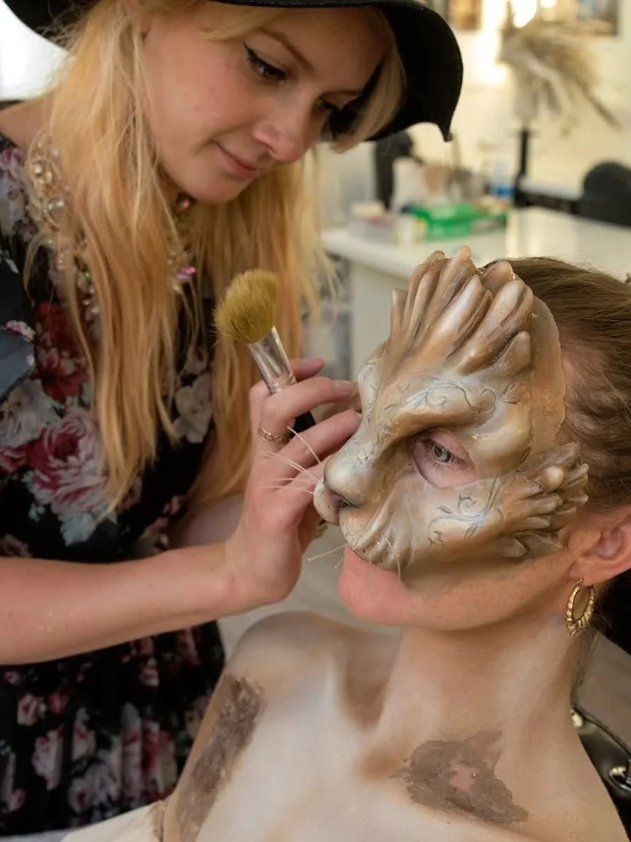 A makeup artist is applying face paint to a woman's face, which is decorated with a lion mask design, in a studio or makeup room.