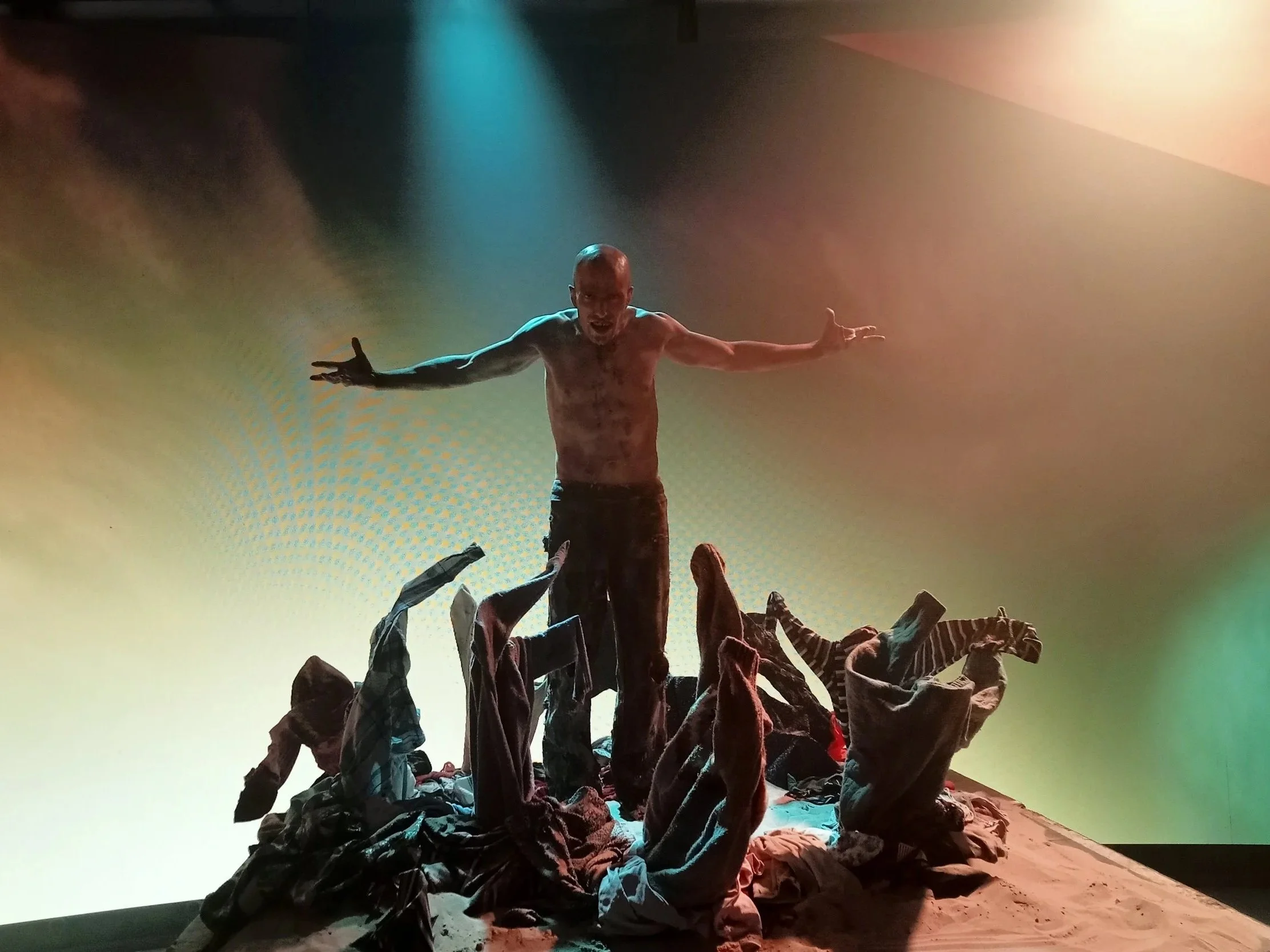 A shirtless man standing on a pile of clothes with arms outstretched, with a colorful backdrop and lighting.