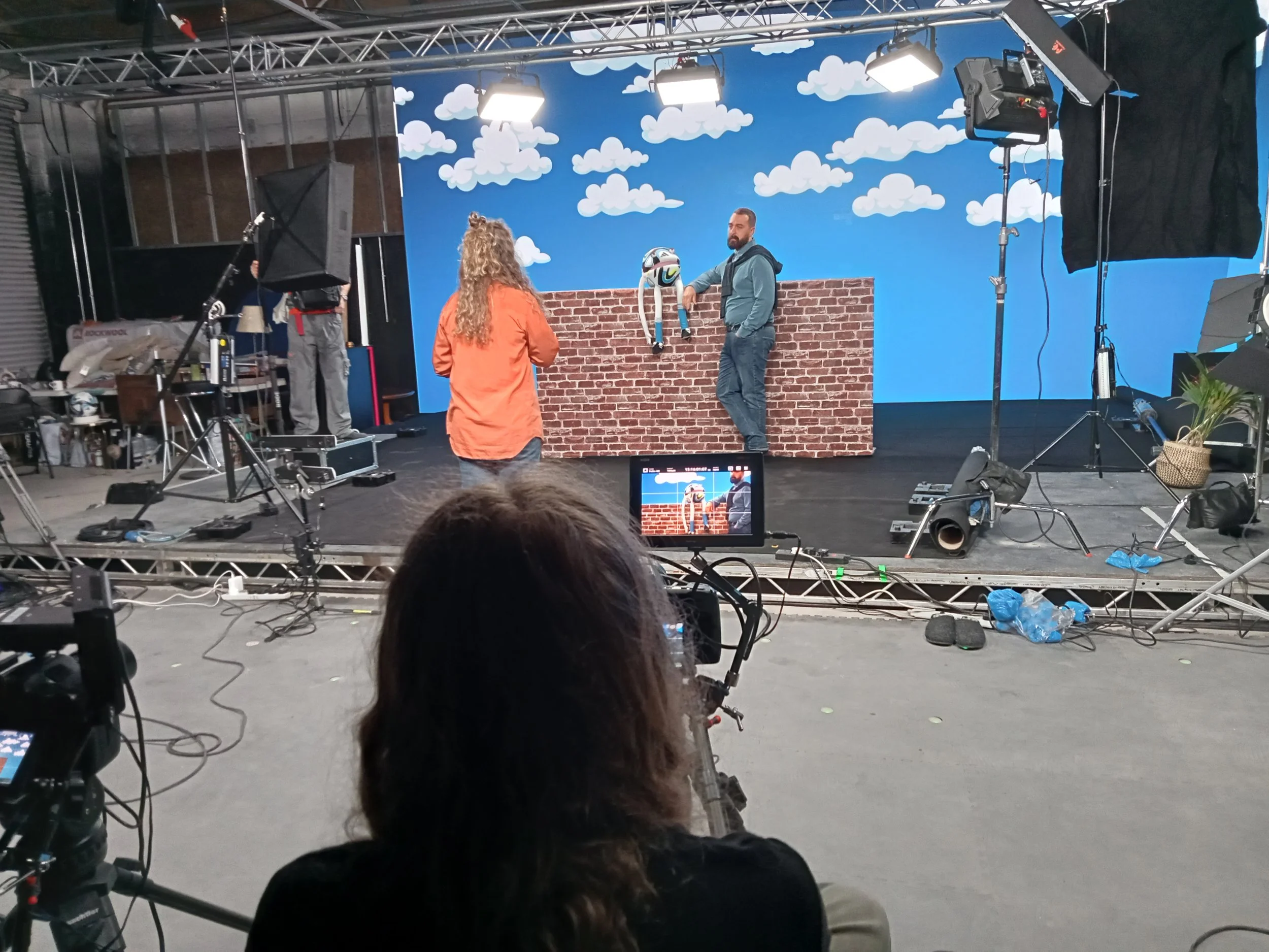 A behind-the-scenes photo of a video shoot or live event setup on a stage with a brick wall backdrop, a blue sky with clouds, and equipment including cameras, lights, and monitors. Two people are on stage, one man holding a helmet and the woman facing away from the camera. There is a person in the foreground operating a camera, capturing the scene.