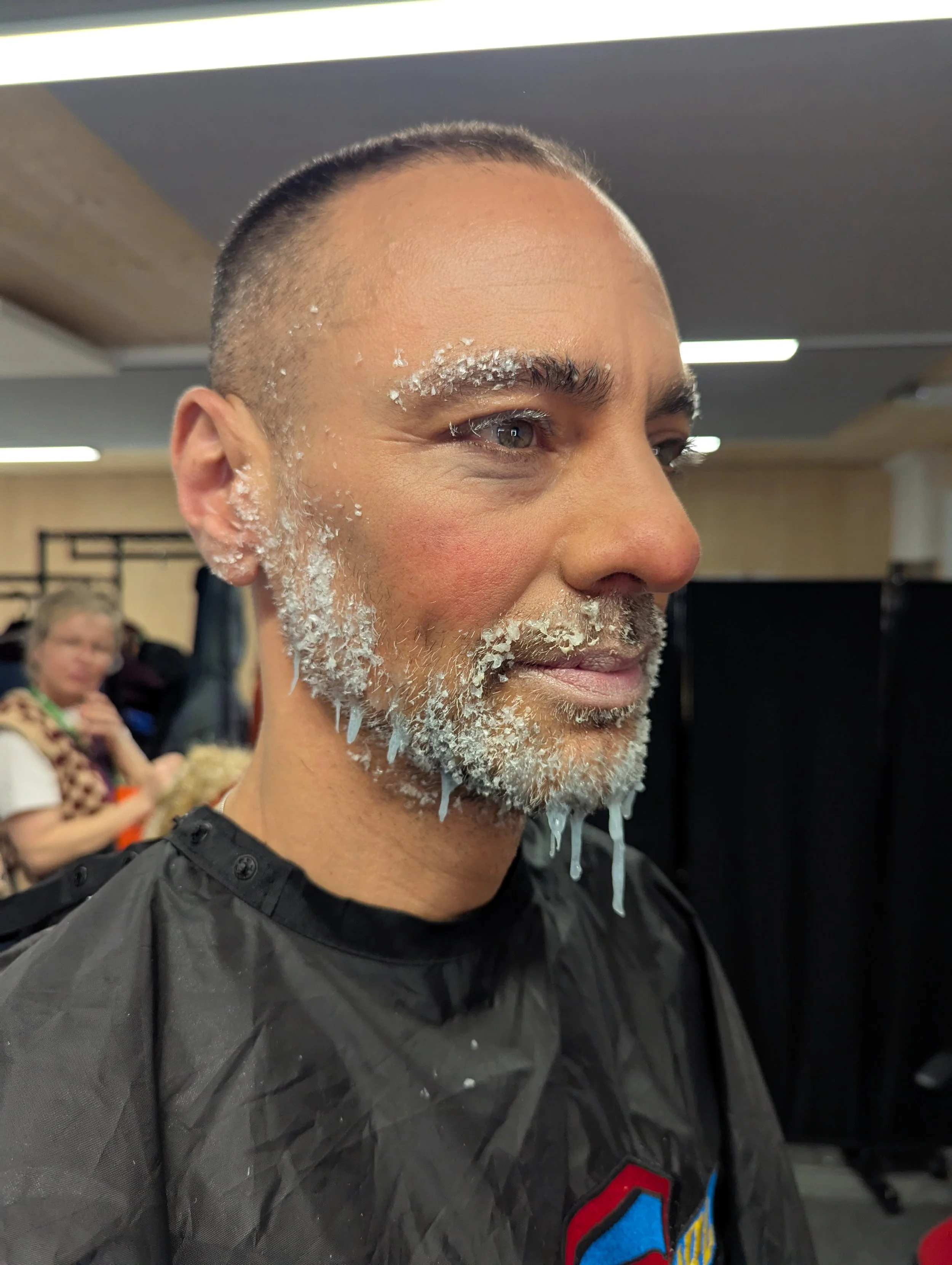 A man with a shaved head and facial hair, covered in white shaving foam or cream on his face, beard, and eyebrows, in a barbershop or salon setting.