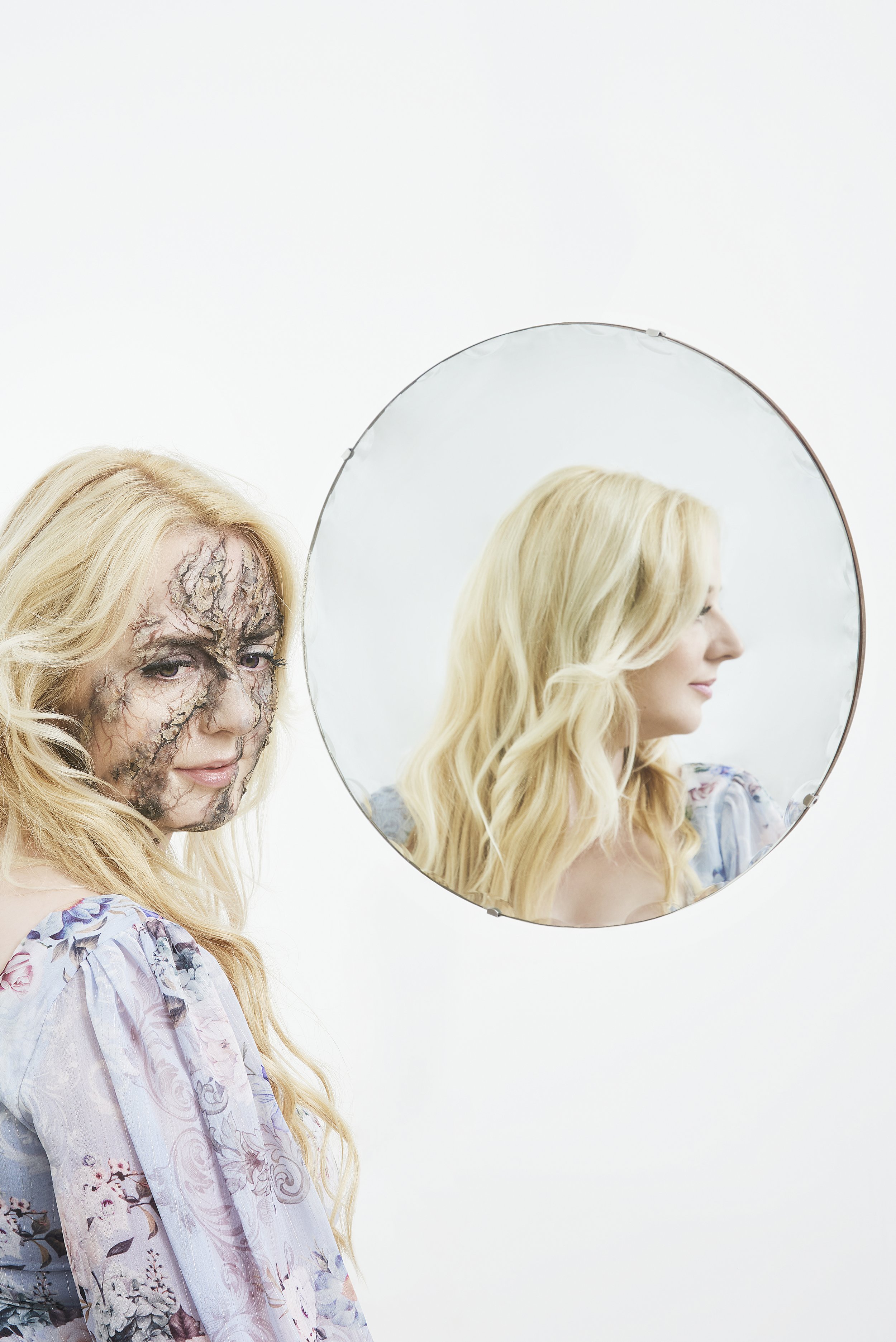 A woman with blonde hair looking into a mirror, her face covered in special effects makeup that makes it look cracked and disfigured.