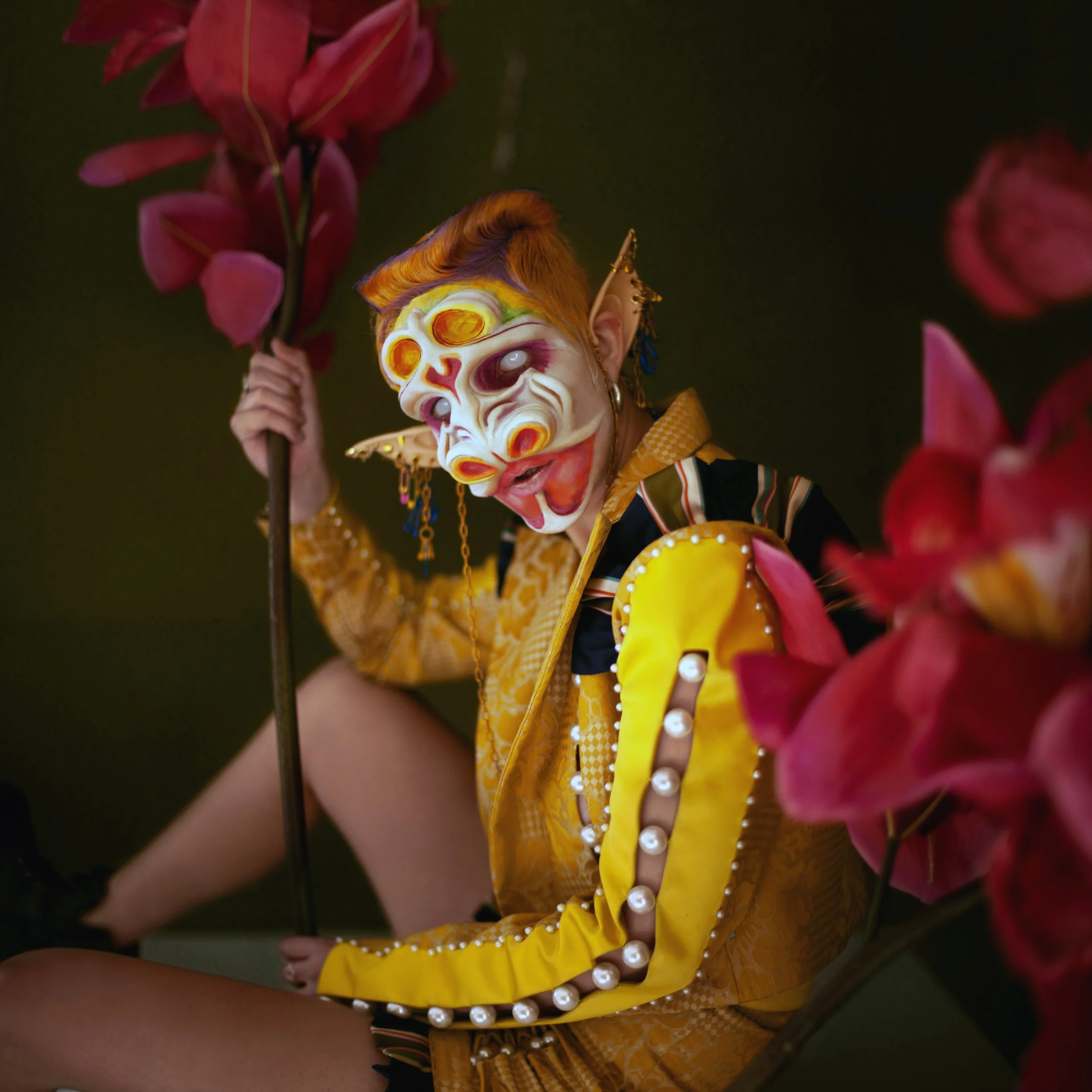 Person wearing a colorful mask with multiple eye designs, pointed ears, and yellow jacket decorated with white pearls, holding a stick, surrounded by pink flowers.
