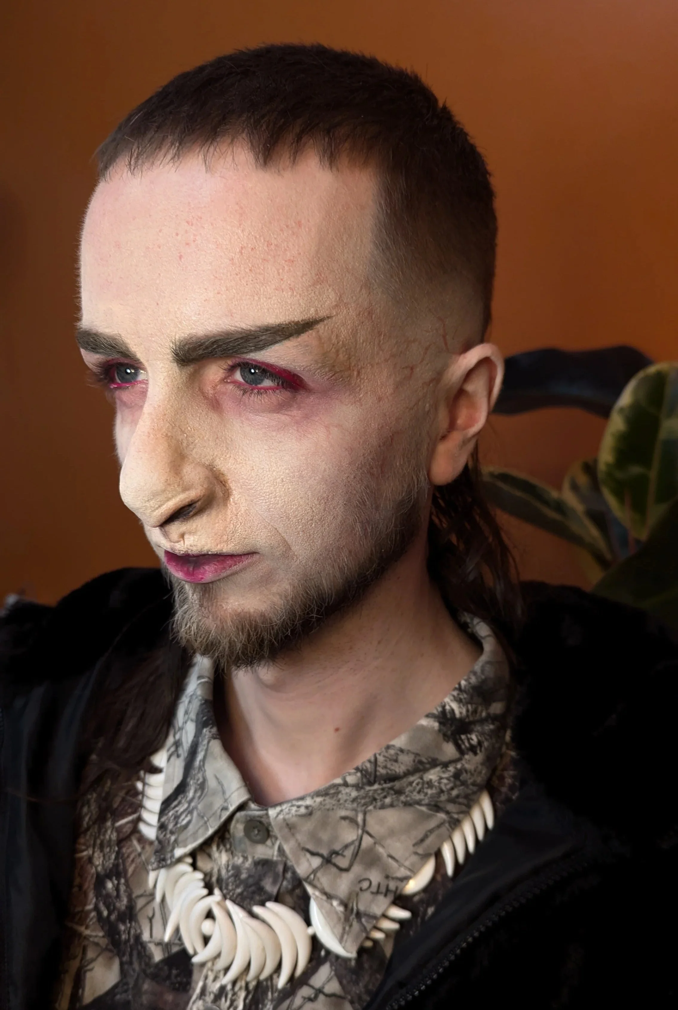 A person with pale makeup, pink eyeshadow, and a styled mohawk hairstyle, wearing a camouflage shirt, a shell necklace, and a black jacket, standing in front of a brown background.