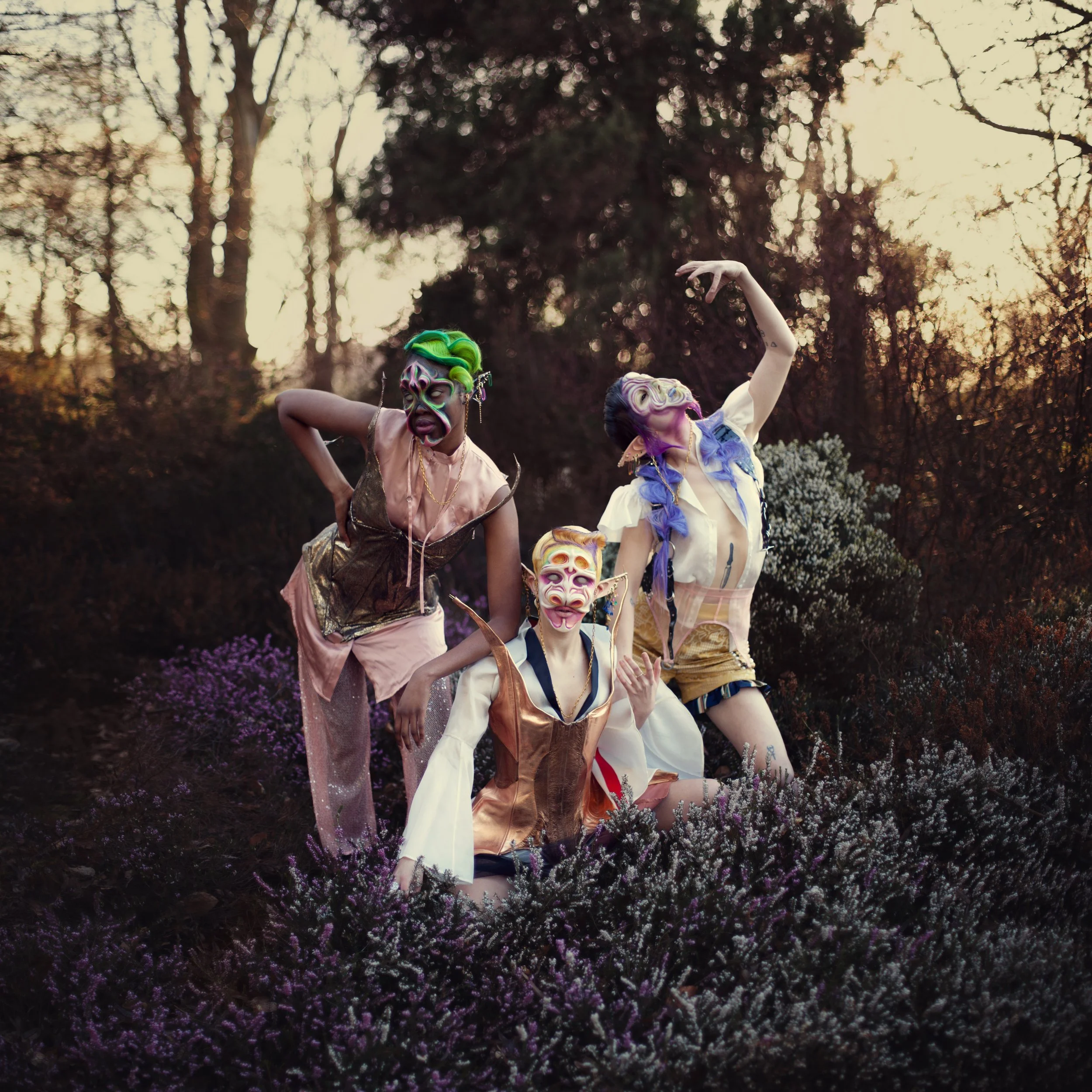 Three individuals in colorful masks and costumes posing outdoors among purple and white flowers during sunset.