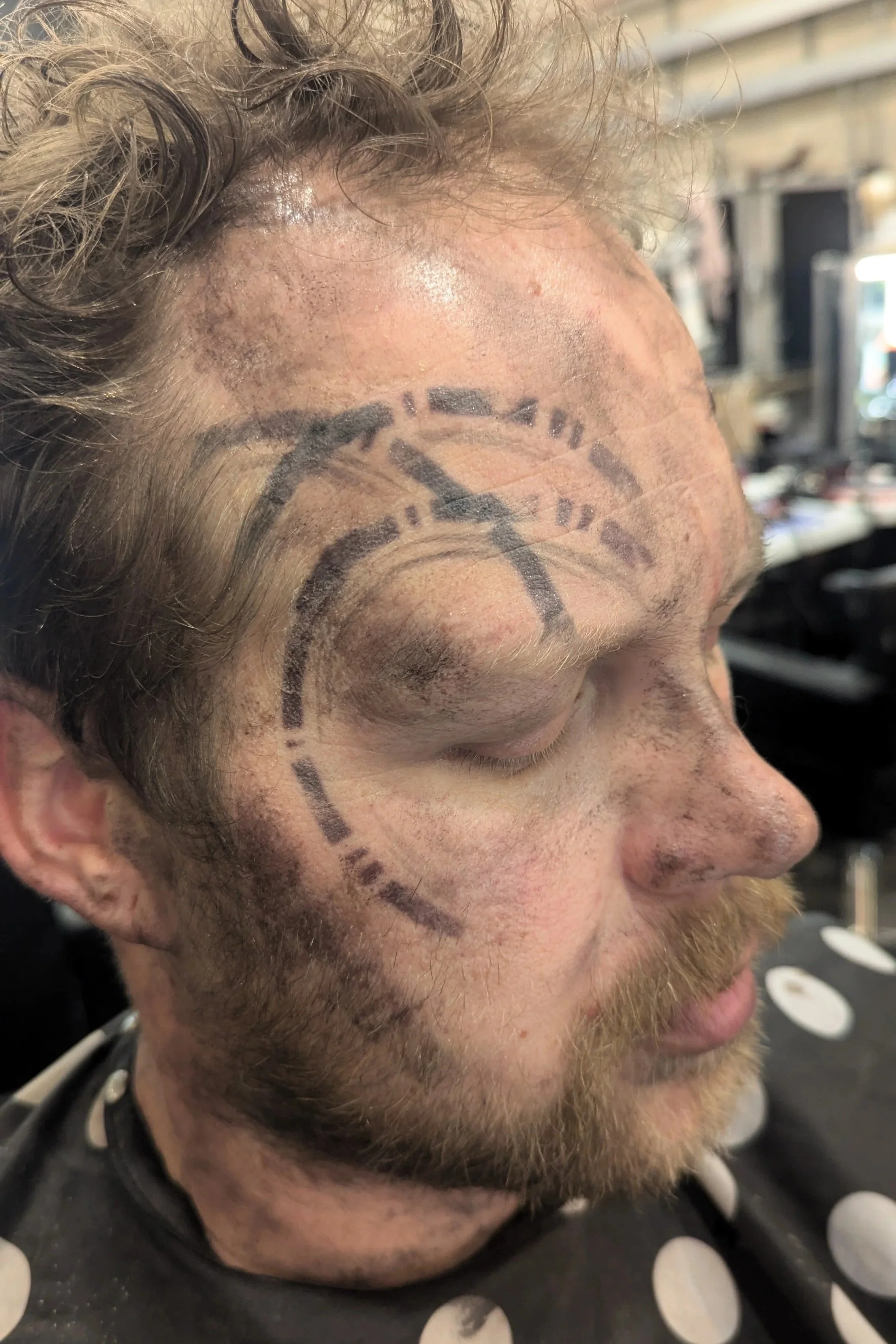 A man with a beard and curly hair has a clock face tattooed on his face, covering his forehead, temple, and cheek.