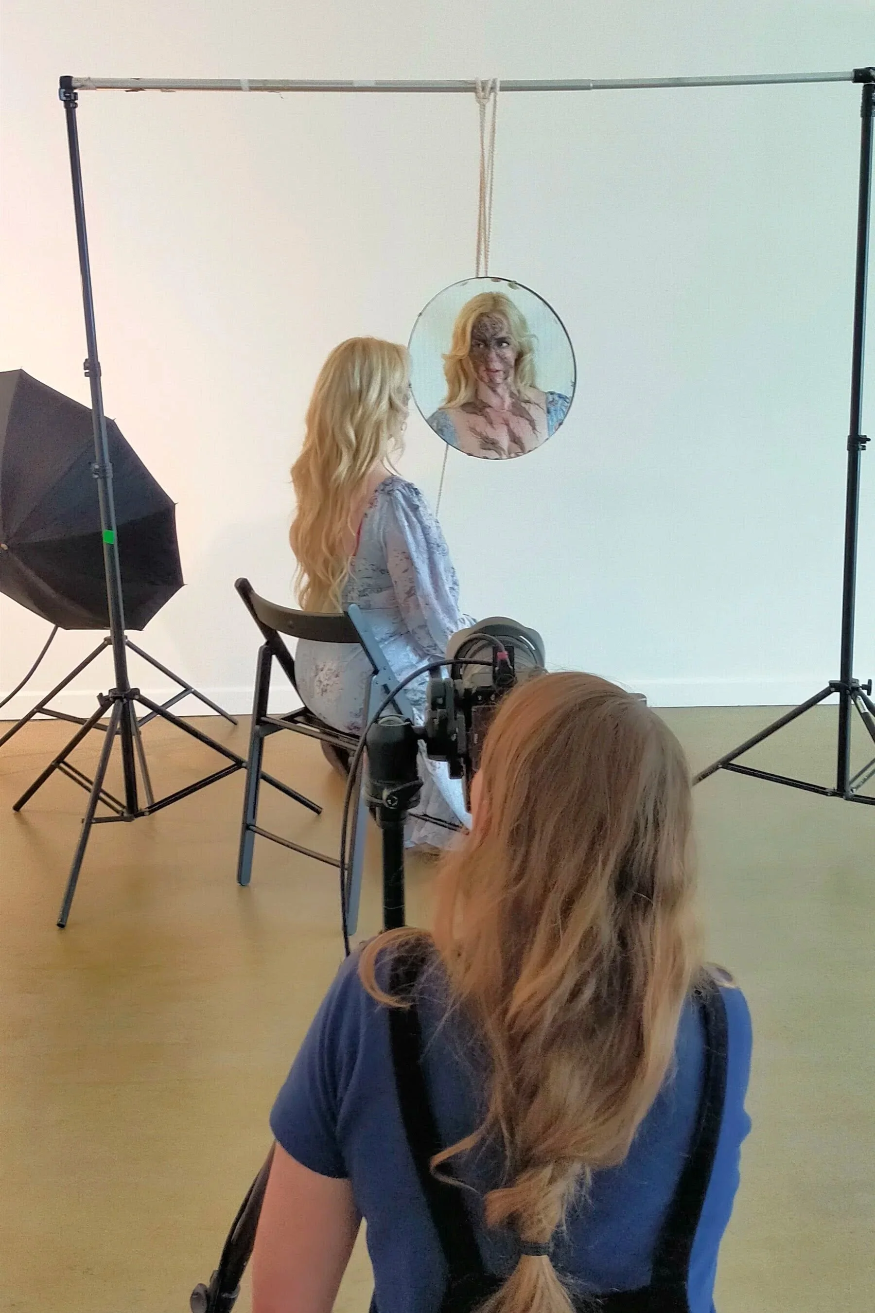 A woman with long blonde hair sitting on a chair, looking at her reflection in a round mirror hung in front of her. A photographer is taking a picture, and a crew member with long hair is operating a camera in the foreground. Studio lighting and equipment are visible.
