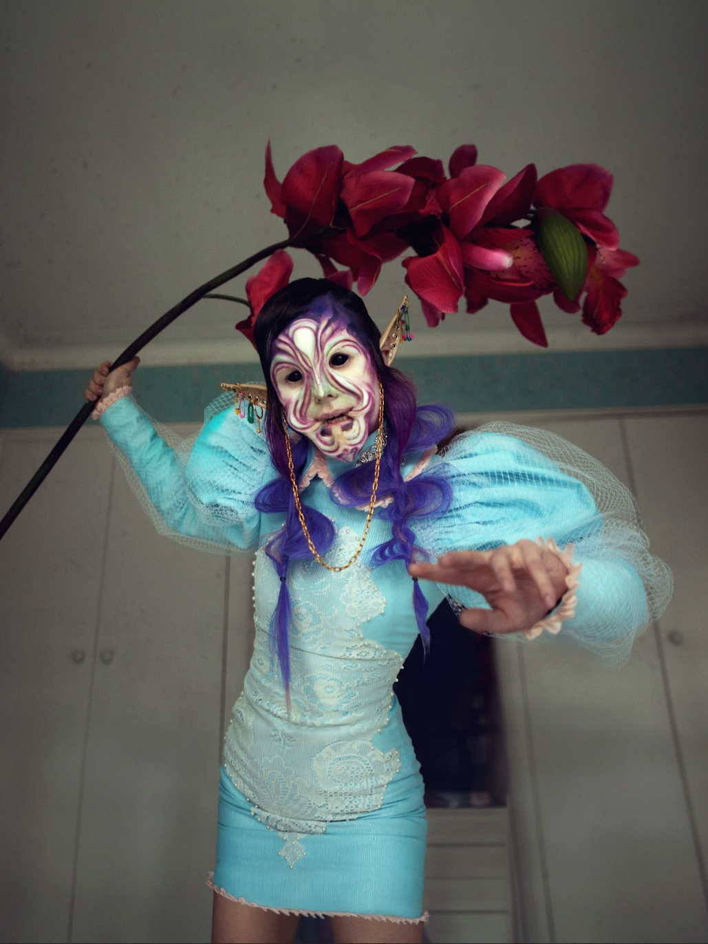 Person in costume with purple braids, colorful makeup, and fantasy accessories holding a large red flower overhead.