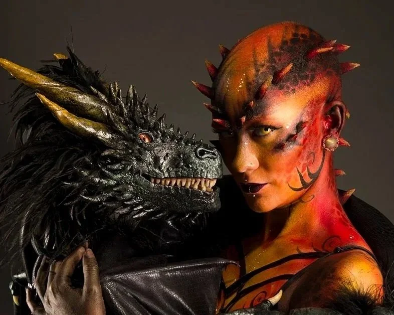 Person dressed as a fiery, dragon-like creature with bright red, orange, and black makeup and costume, posing with a person wearing a detailed, black dragon or monster mask with horns and sharp teeth.