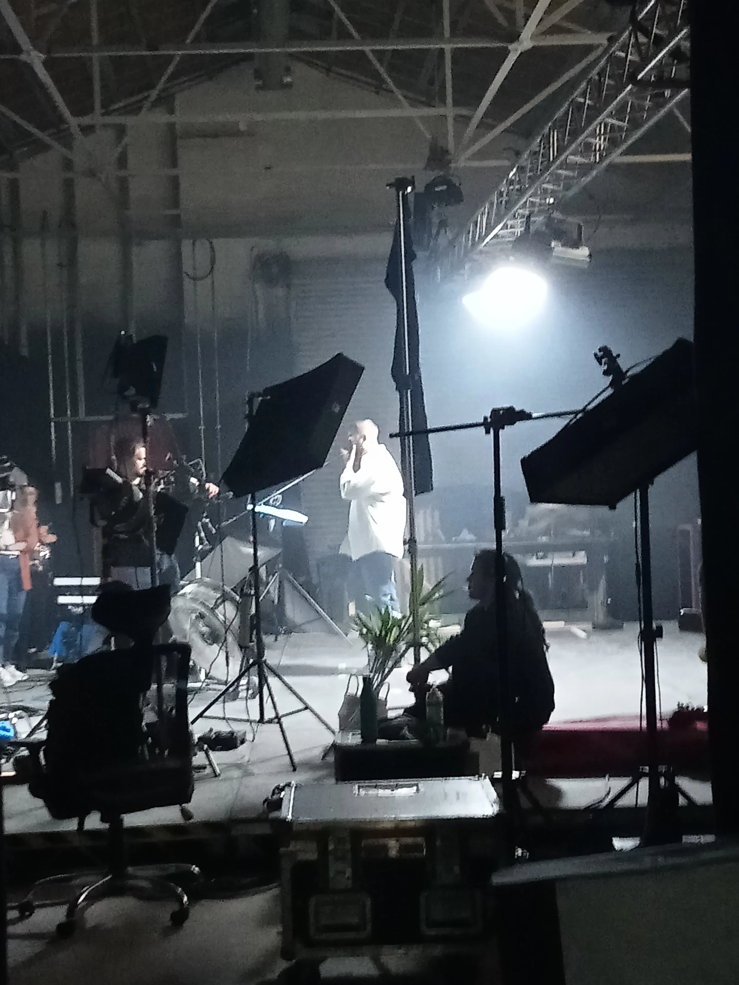 Behind the scenes view of a music or film set with equipment and lighting. A person in a white jacket is talking on a phone or microphone, with crew members and camera equipment around.