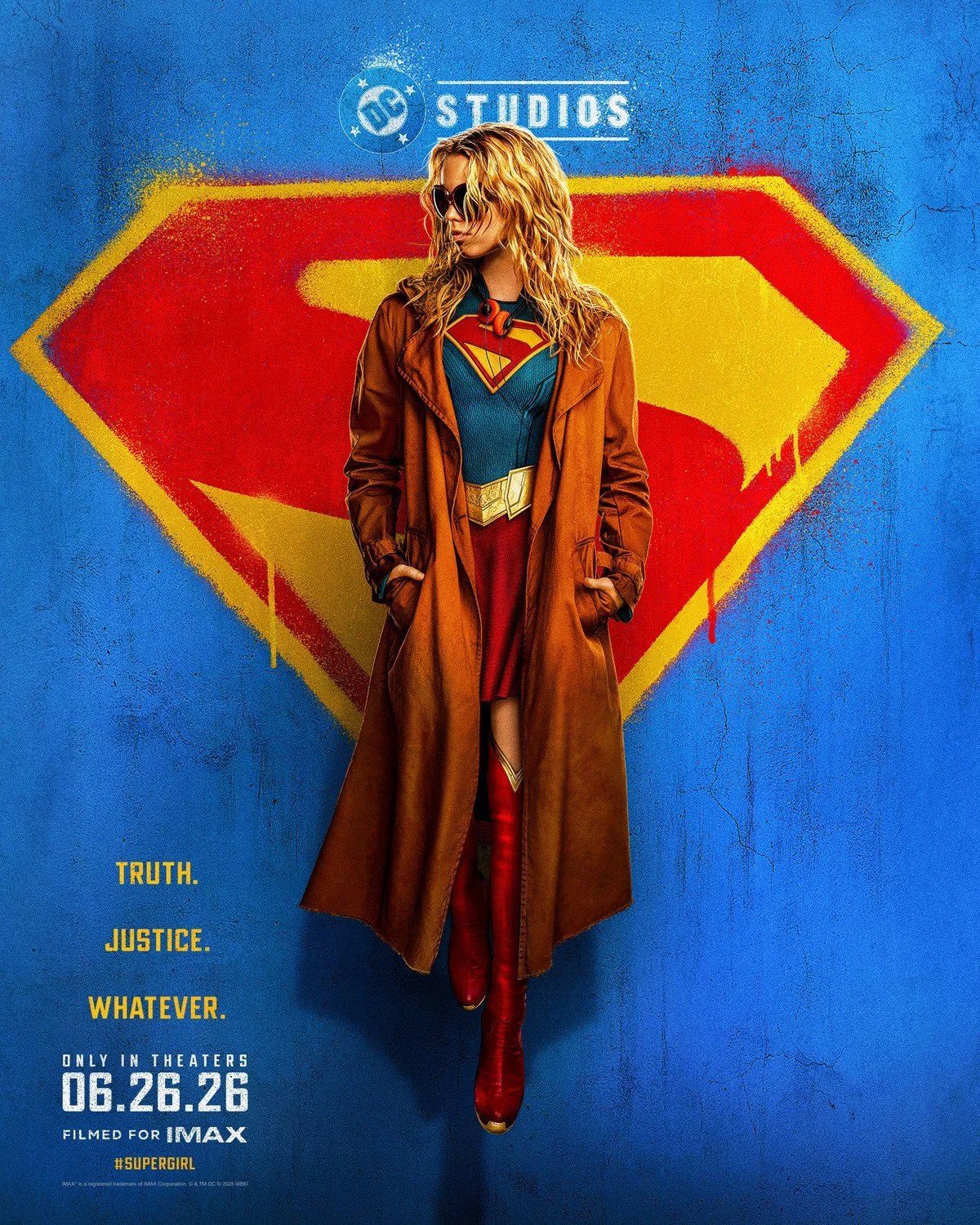 Poster of a woman dressed as Supergirl with a long brown coat and red boots, standing in front of a wall with a large Supergirl emblem, wearing sunglasses and a blue suit with the Supergirl logo.