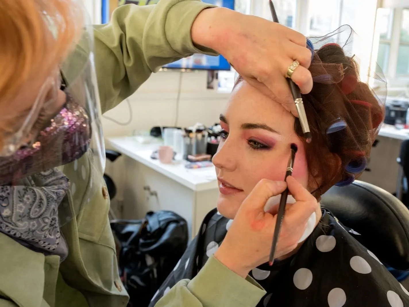 Makeup artist applying eyeshadow to a woman's eyelids in a brightly lit room.