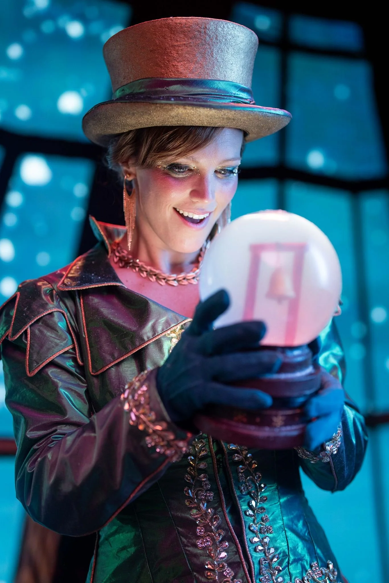 A woman wearing a shiny, patterned jacket, black gloves, and a large, metallic top hat looks at a glowing orb with a transparent rectangular shape inside, in a brightly lit, futuristic setting.