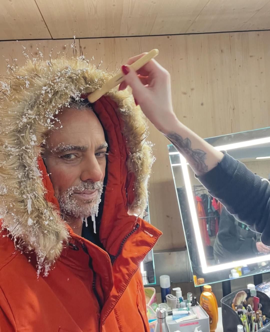 Person with snowy snow on face and eyebrows, wearing a thick orange parka with a furry hood, getting makeup or costume adjustments done in front of a mirror with bright lights inside a wooden room.