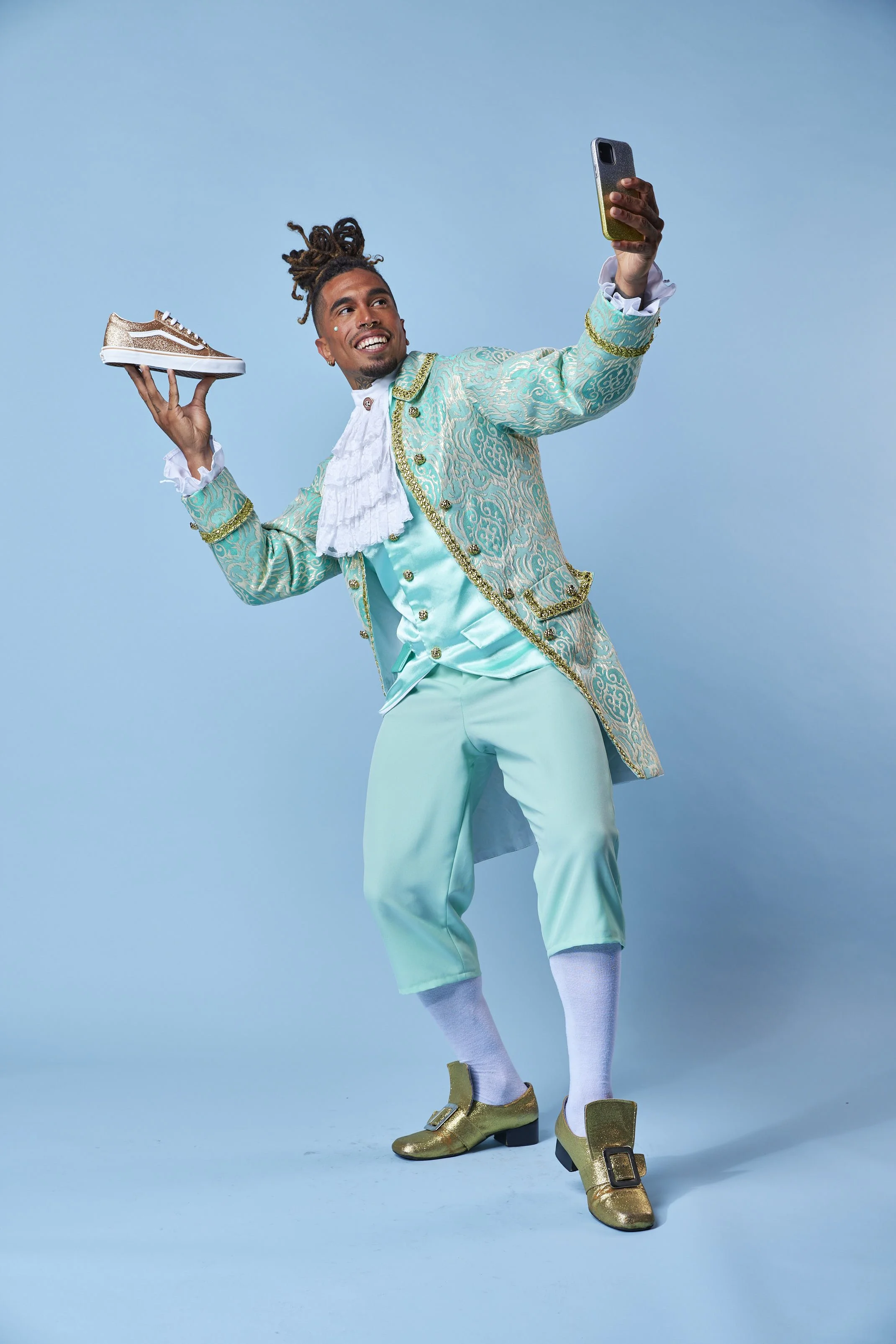 Person dressed in an ornate, historical costume taking a selfie with a smartphone, holding a sneaker in the other hand, against a blue background.