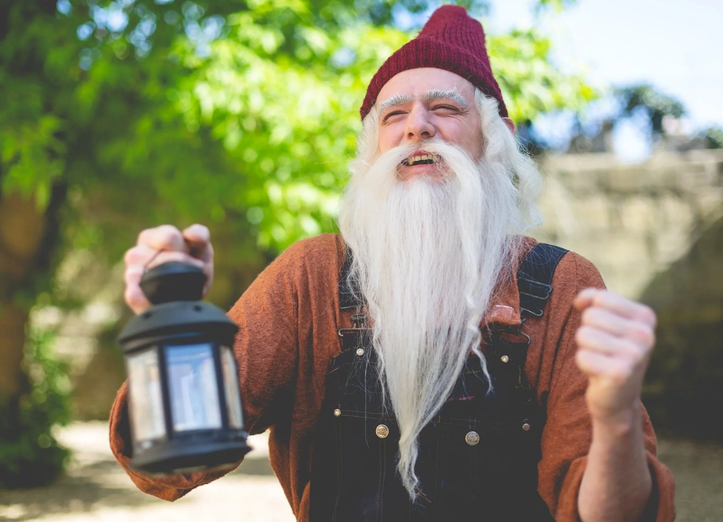 An elderly man with a long white beard and white hair, wearing a red beanie, smiling and holding a lantern outdoors in a wooded area.