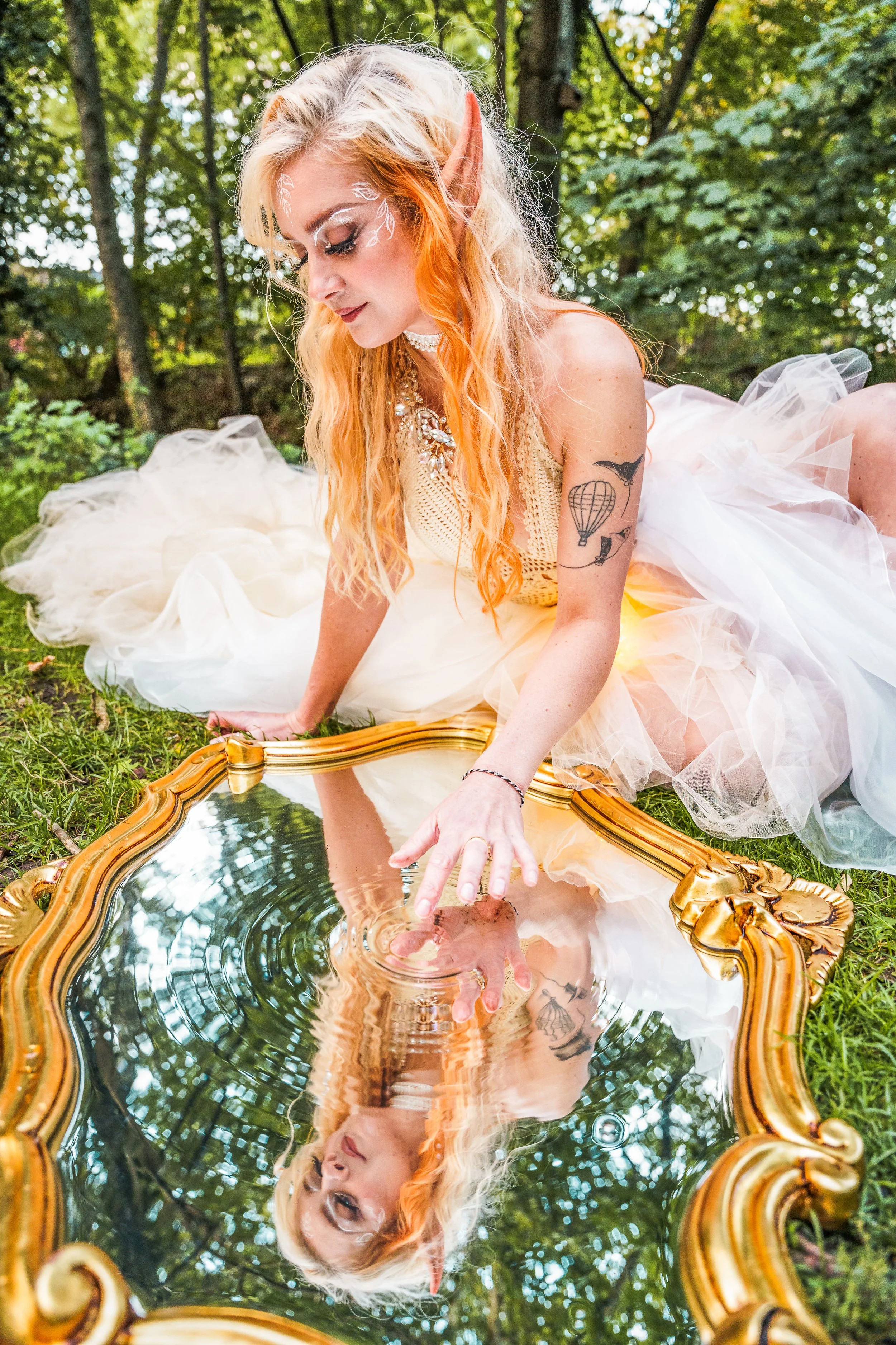 A woman with elf ears and orange hair in a fairy costume, sitting on grass outdoors, touching a decorative mirror laid on the ground reflecting her face.