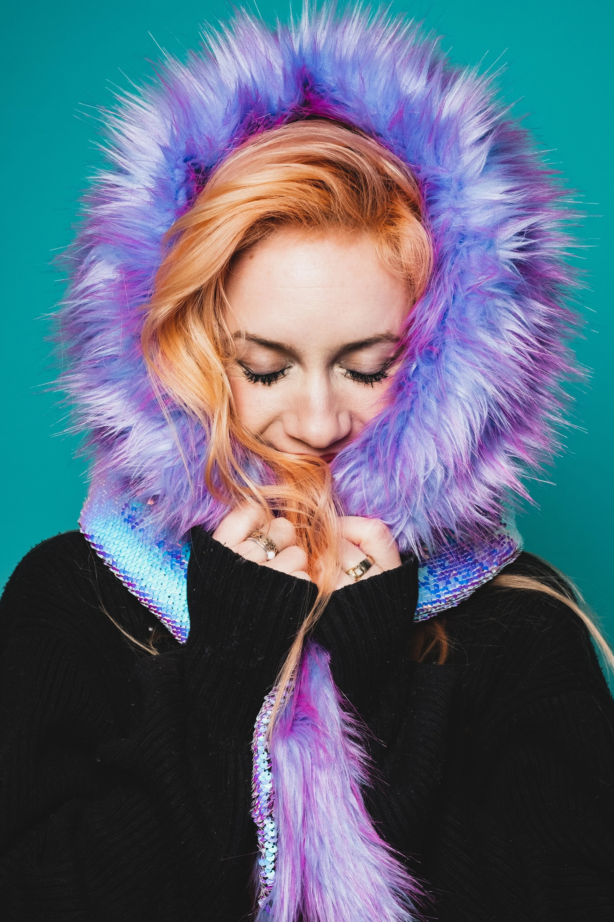 A woman with pink hair wearing a black top and a purple furry hood with iridescent details, looking down with a gentle smile.