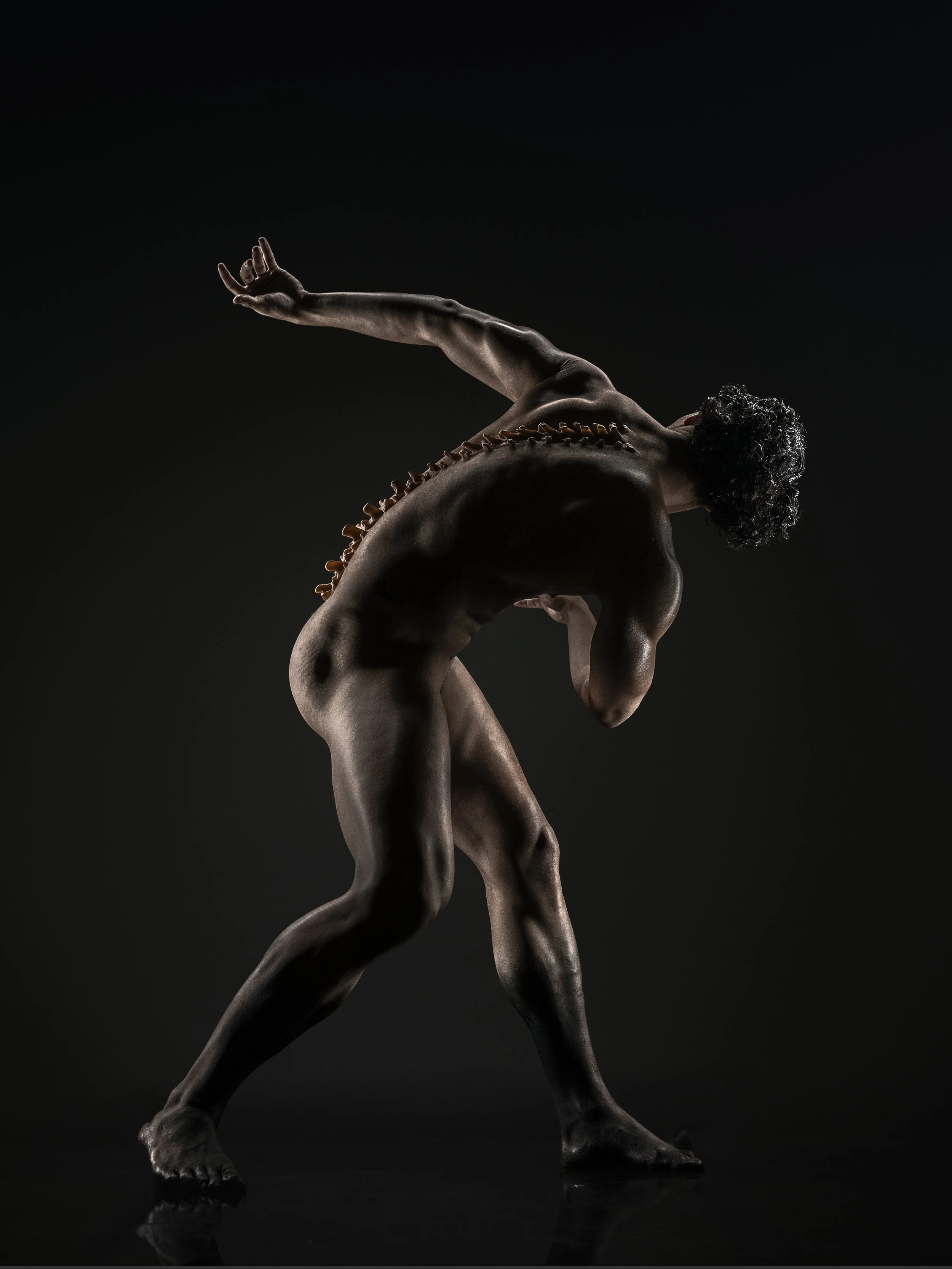 A male dancer with dark curly hair performs a contemporary dance pose against a black background, with his body bent and arms extended.