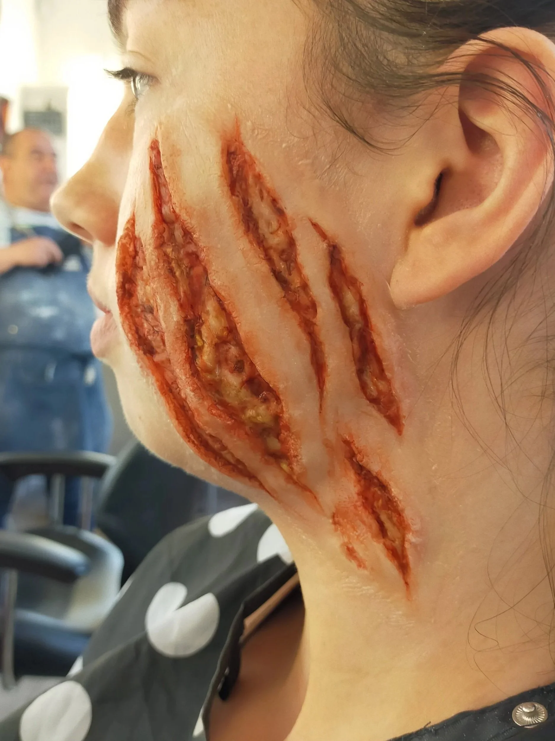 A woman with realistic-looking fake scars and wounds on her cheek, resembling a horror or Halloween makeup effect.