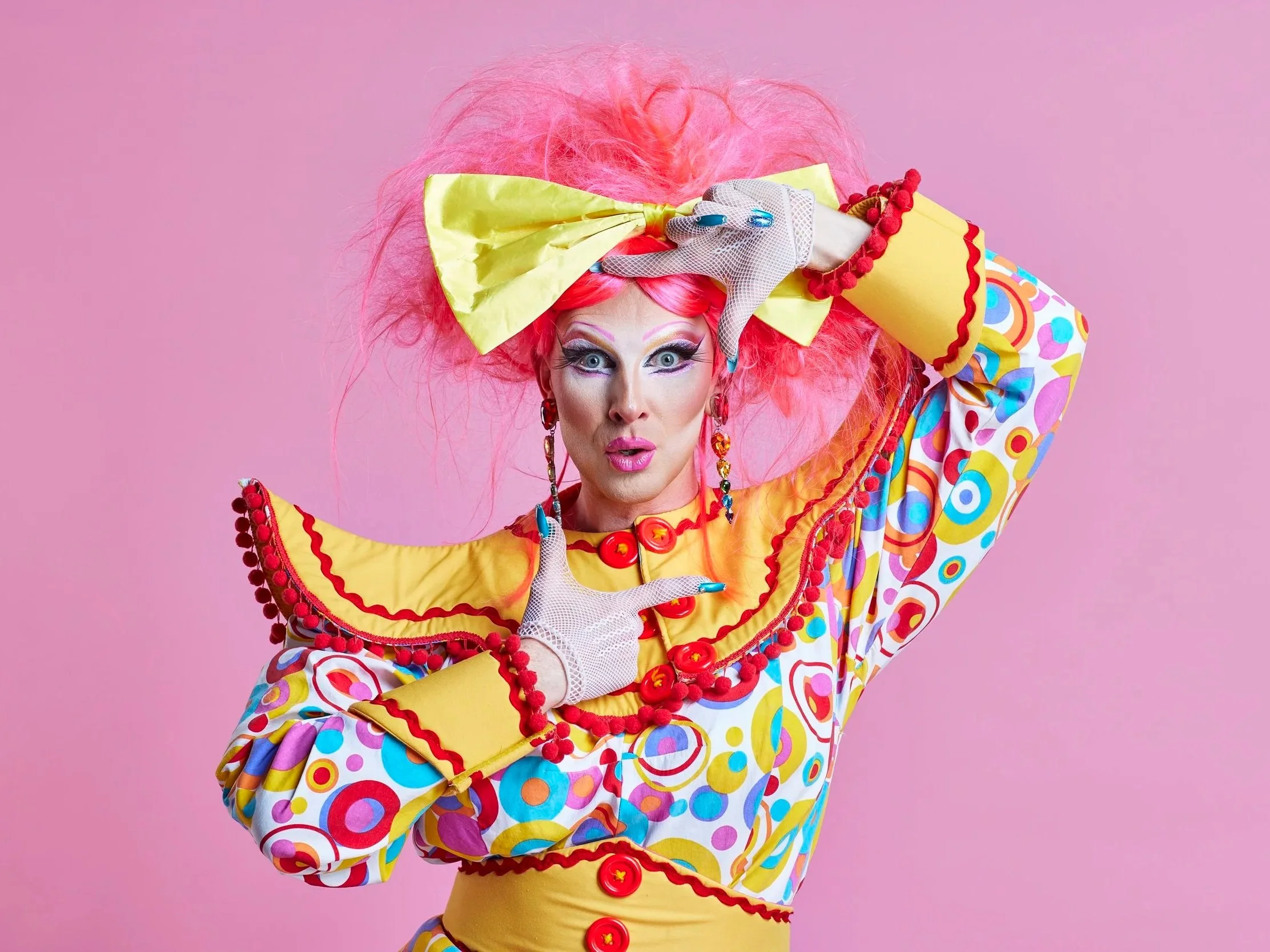 A person dressed in vibrant, colorful clown costume with pink hair, bold makeup, and playful accessories, posing against a pink background.