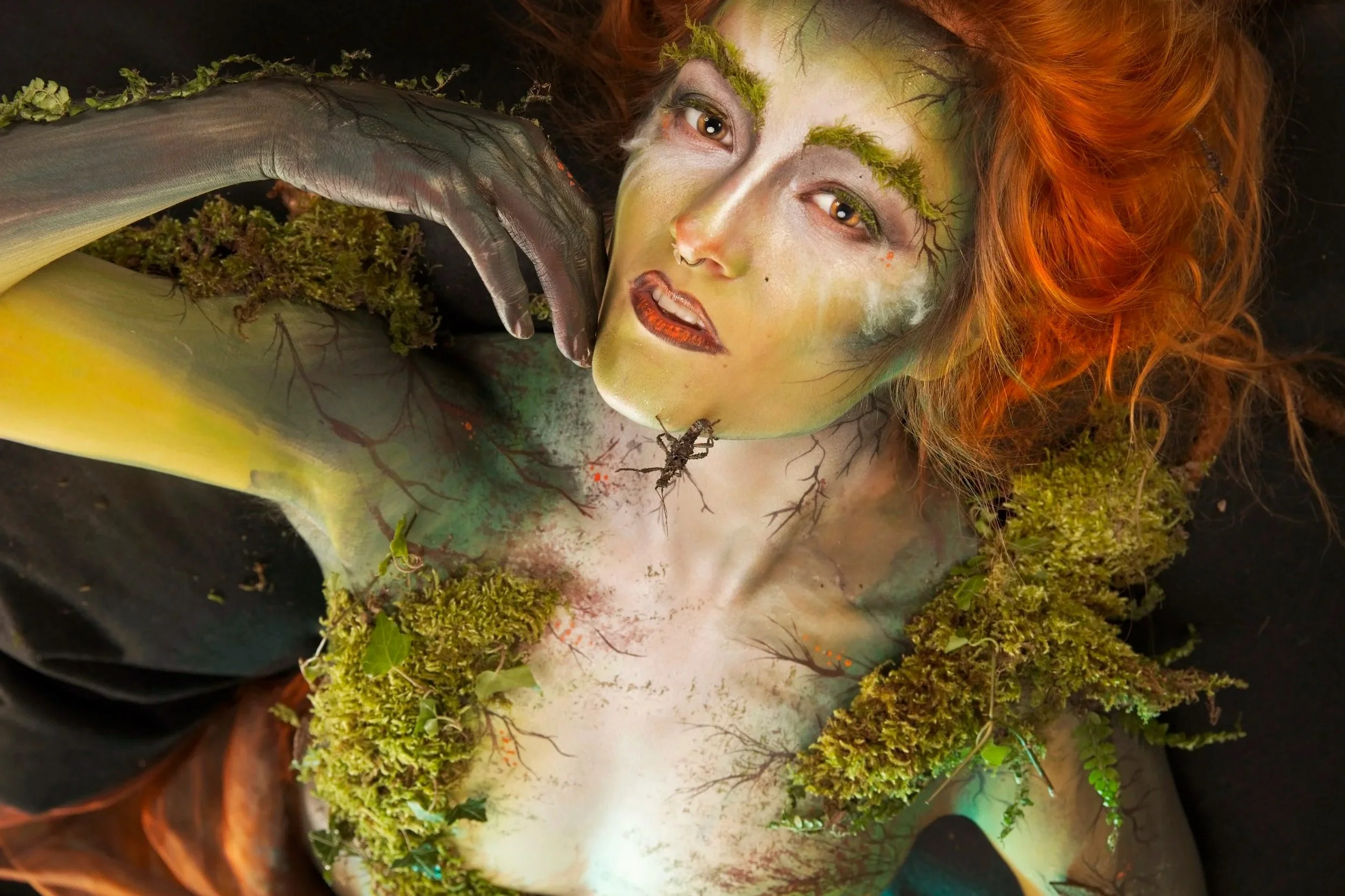 A person is lying down with body and face painted to resemble a forest scene. The body paint includes tree branches and moss, with real moss and leaves attached to the body. The person's face is painted with green, brown, and white colors, with makeup mimicking tree bark, and they have wild, curly red hair.