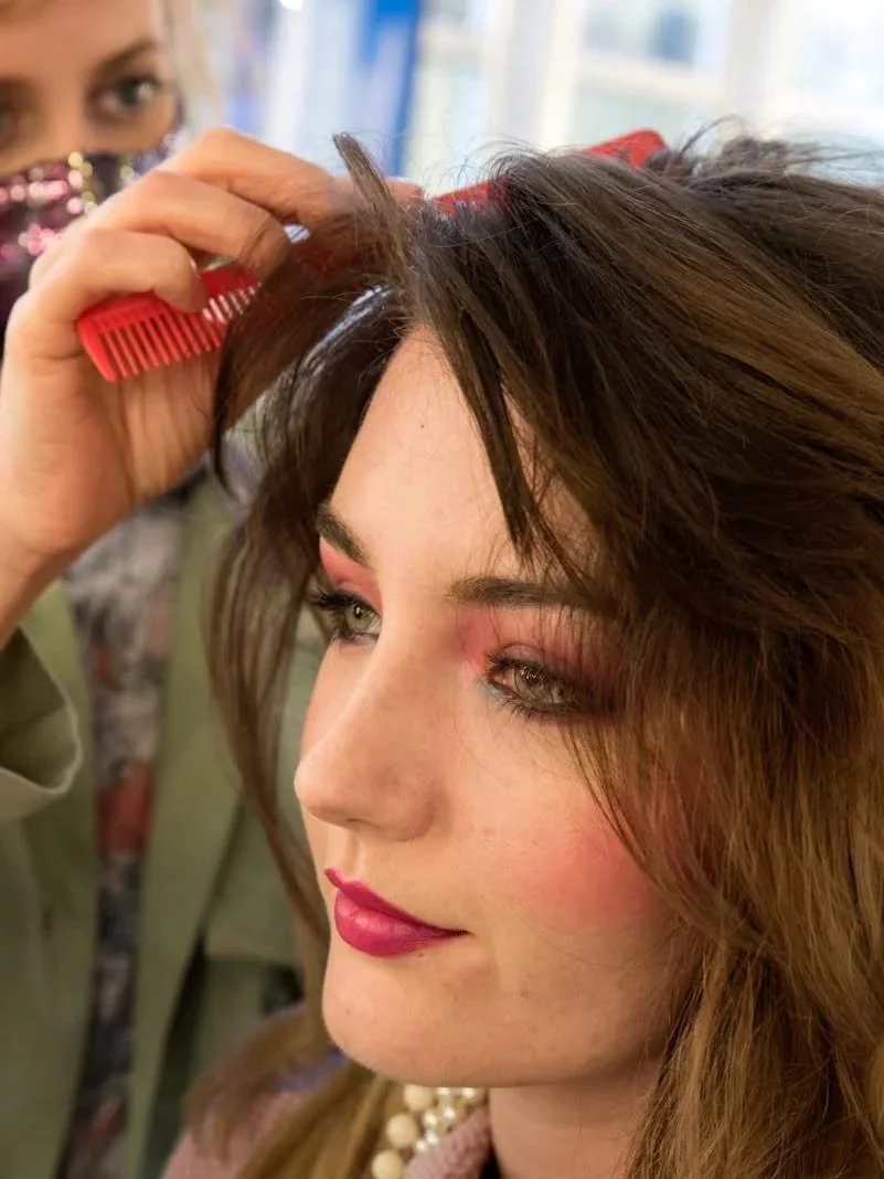 A woman with styled brown hair and makeup is getting her hair cut by a stylist using a red comb at a salon.