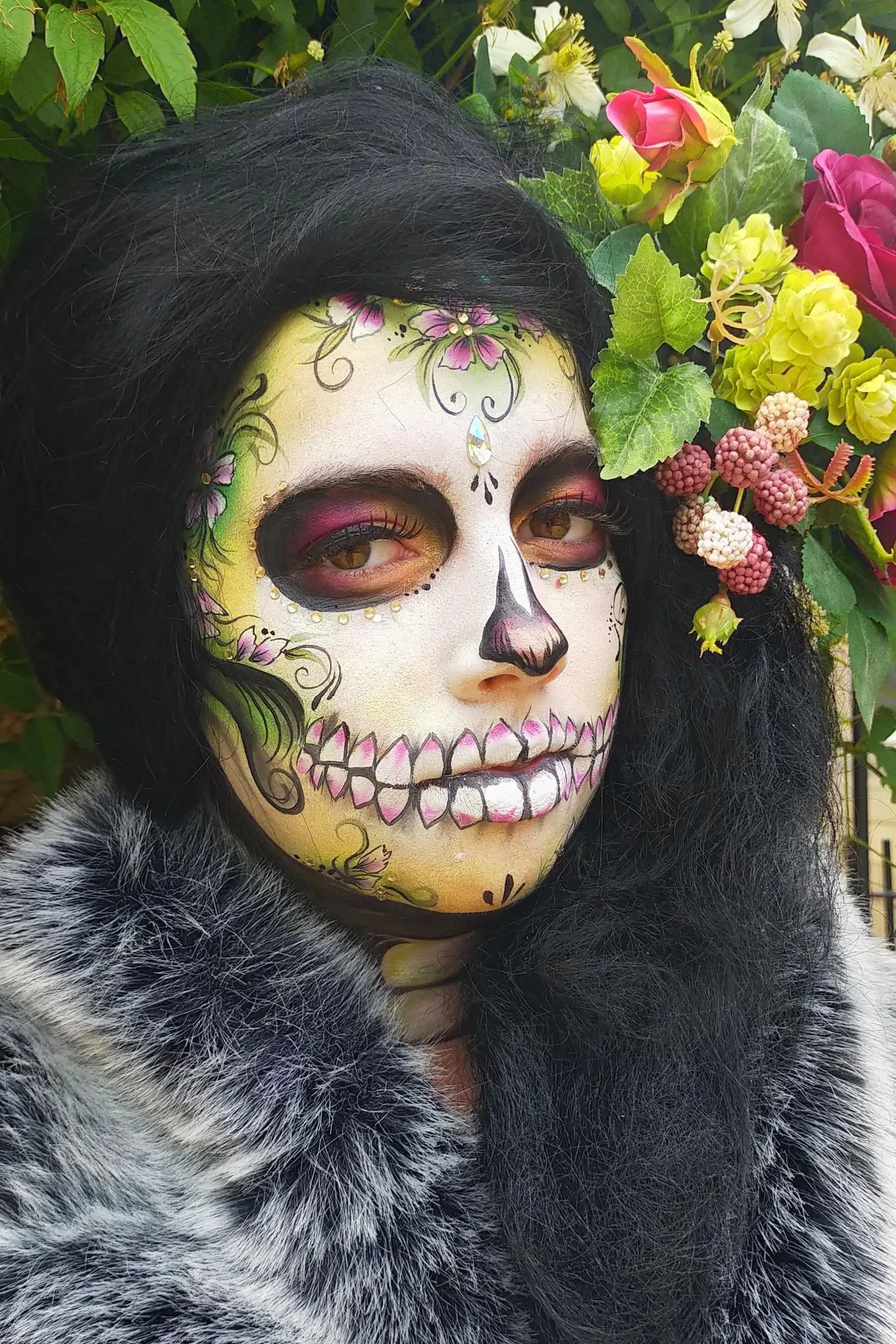 Person with sugar skull makeup and Day of the Dead decorations, surrounded by colorful flowers and wearing a fur coat.