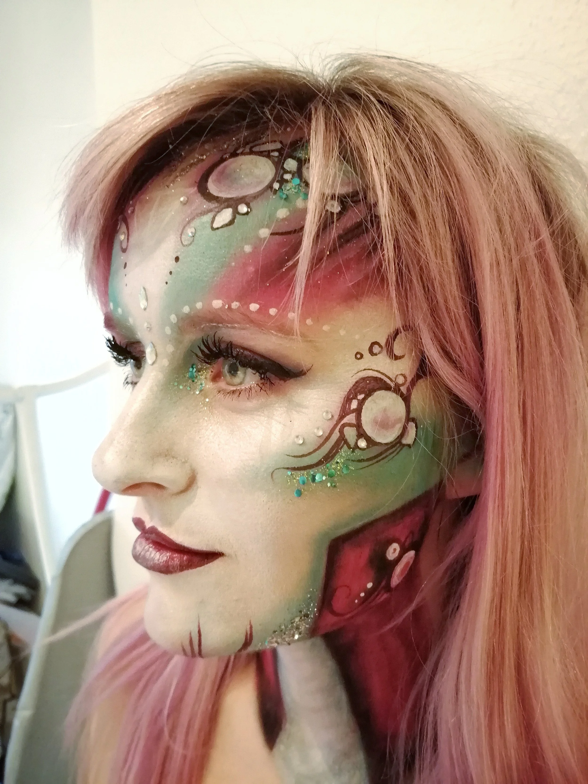 Close-up of a woman with elaborate, colorful fantasy makeup featuring bubbles, swirls, and glitter, with pink hair.