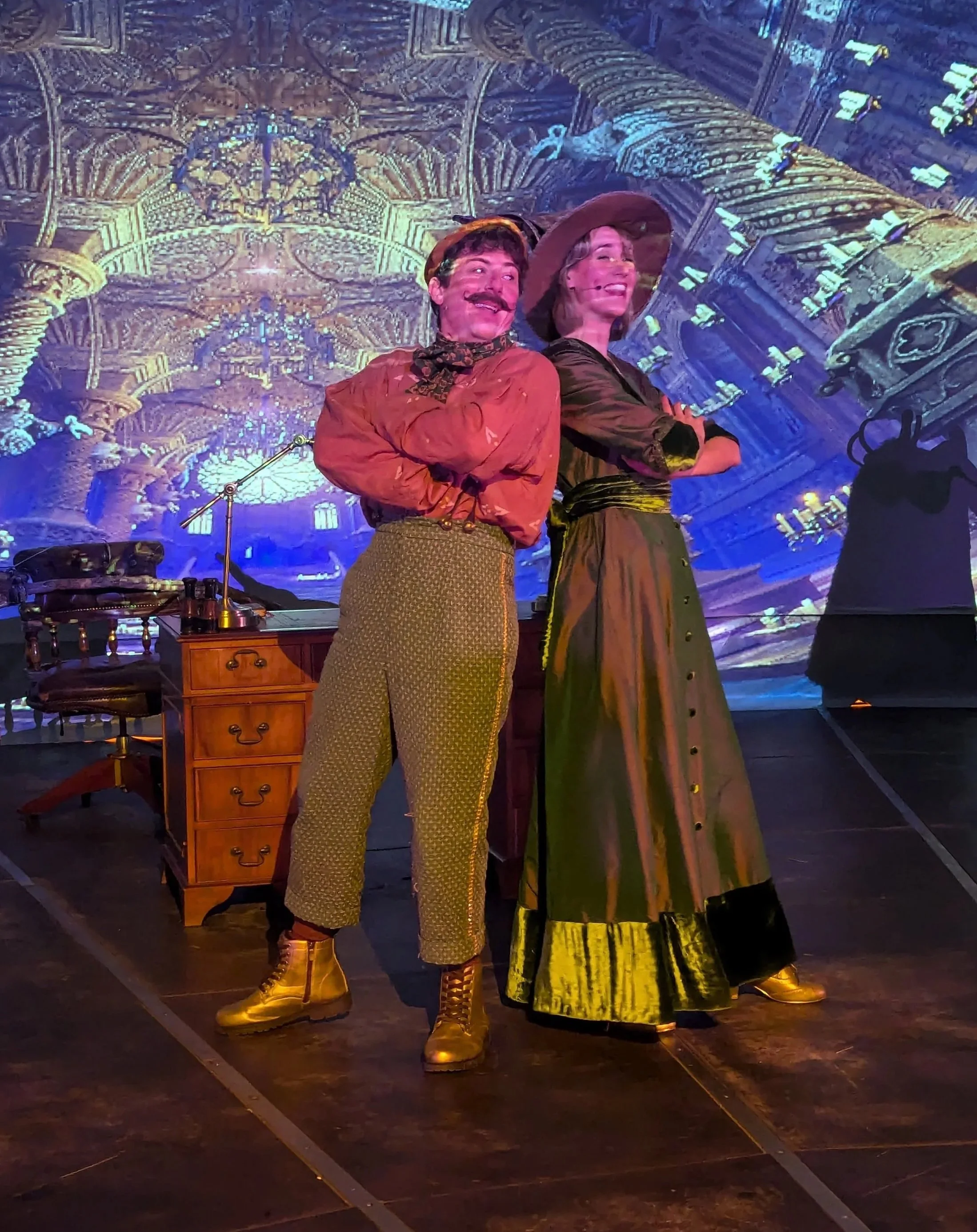 Two performers dressed in period costumes, standing with arms crossed on stage with a detailed, ornate background projection.