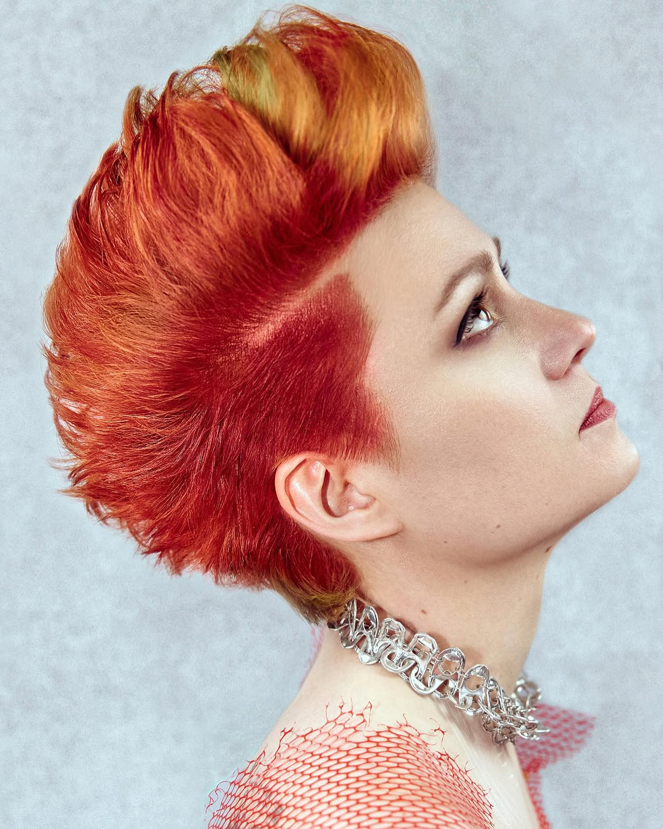A woman with a vibrant red and orange mohawk hairstyle, wearing a metallic chain necklace, and red fishnet clothing visible on her shoulders.