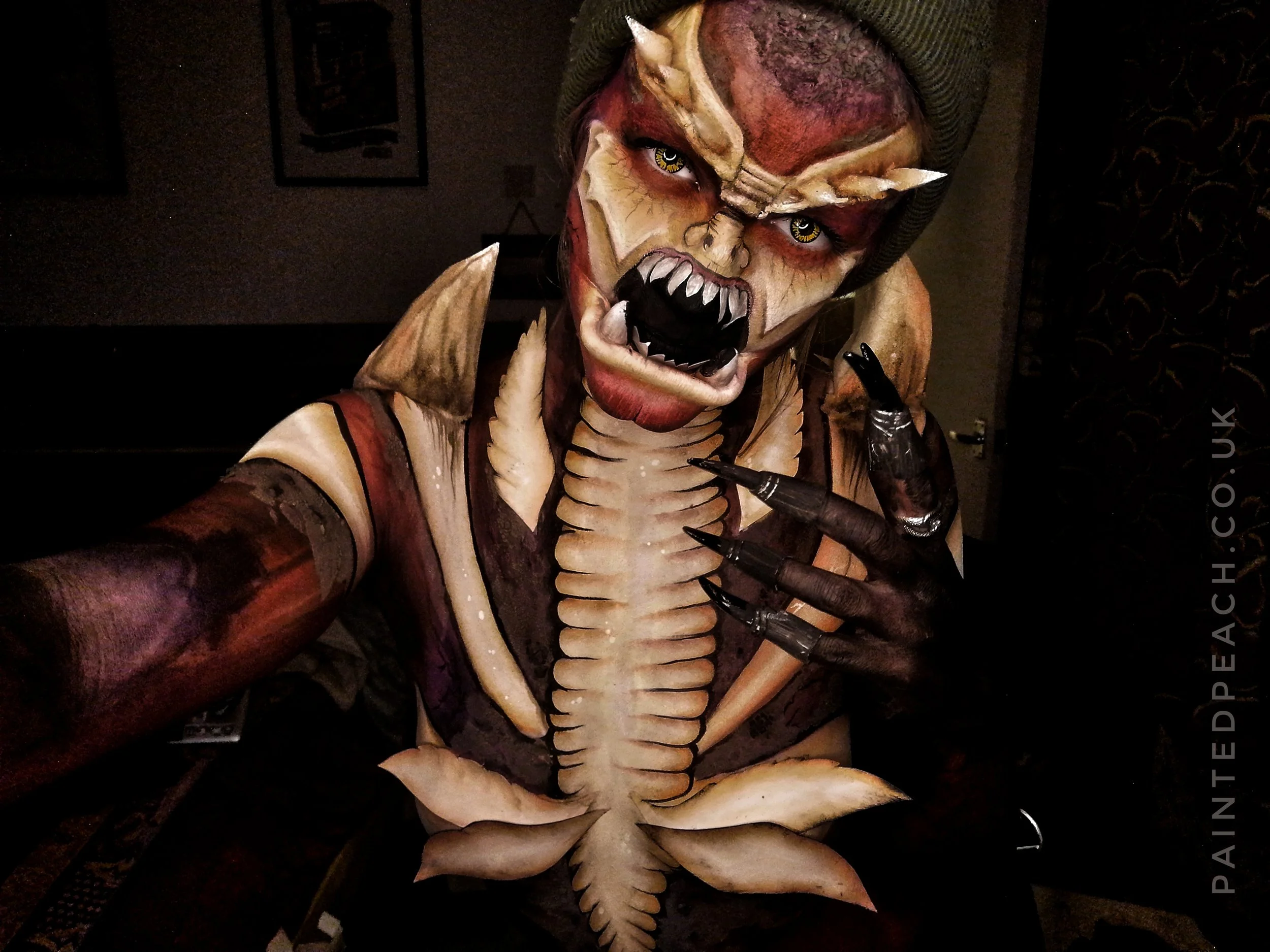 A person dressed in detailed terrifying monster costume with sharp teeth, claws, and exaggerated facial features taking a selfie in a dim room.