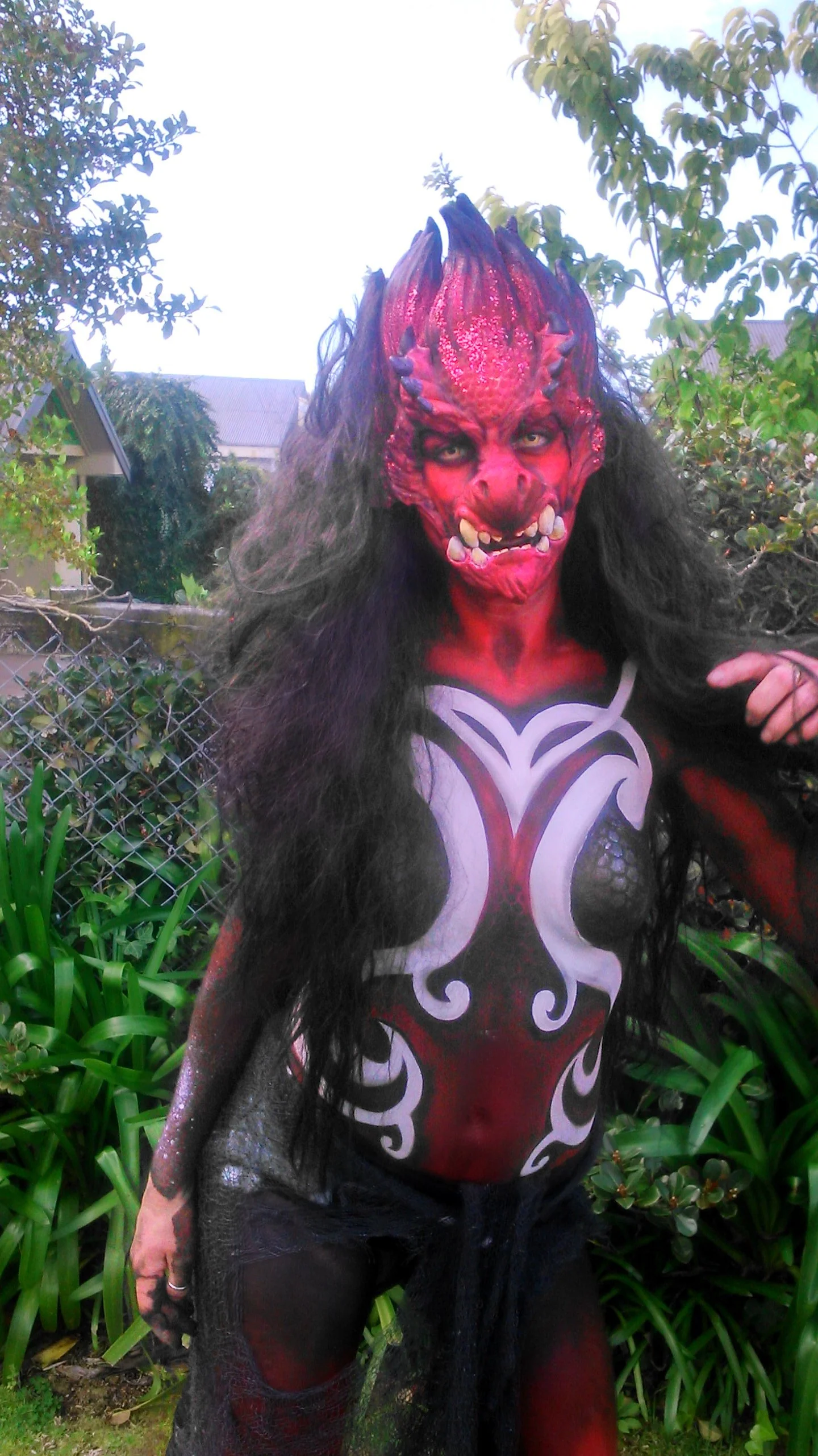 Person dressed as a fantasy creature with red, black, and white body paint, wearing a puppet mask with horns, sharp teeth, and long dark hair, standing outdoors among greenery.