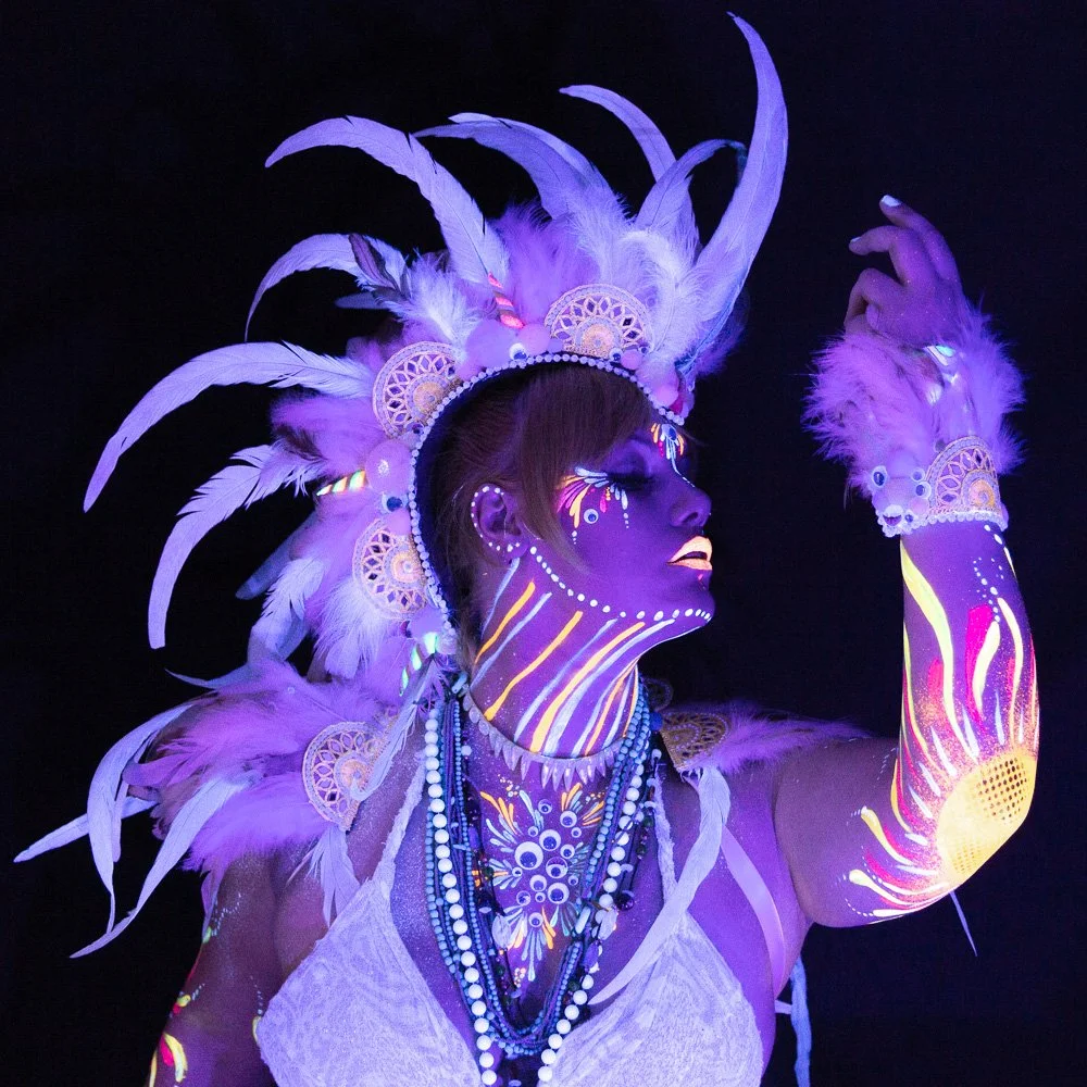 Person with elaborate feather headdress and body paint illuminated by blacklight.