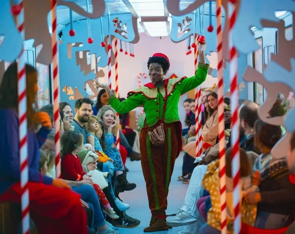 A performer dressed as an elf in green and red costume entertains an audience, including children and adults, at a festive event decorated with holiday-themed cutouts and candy cane props.