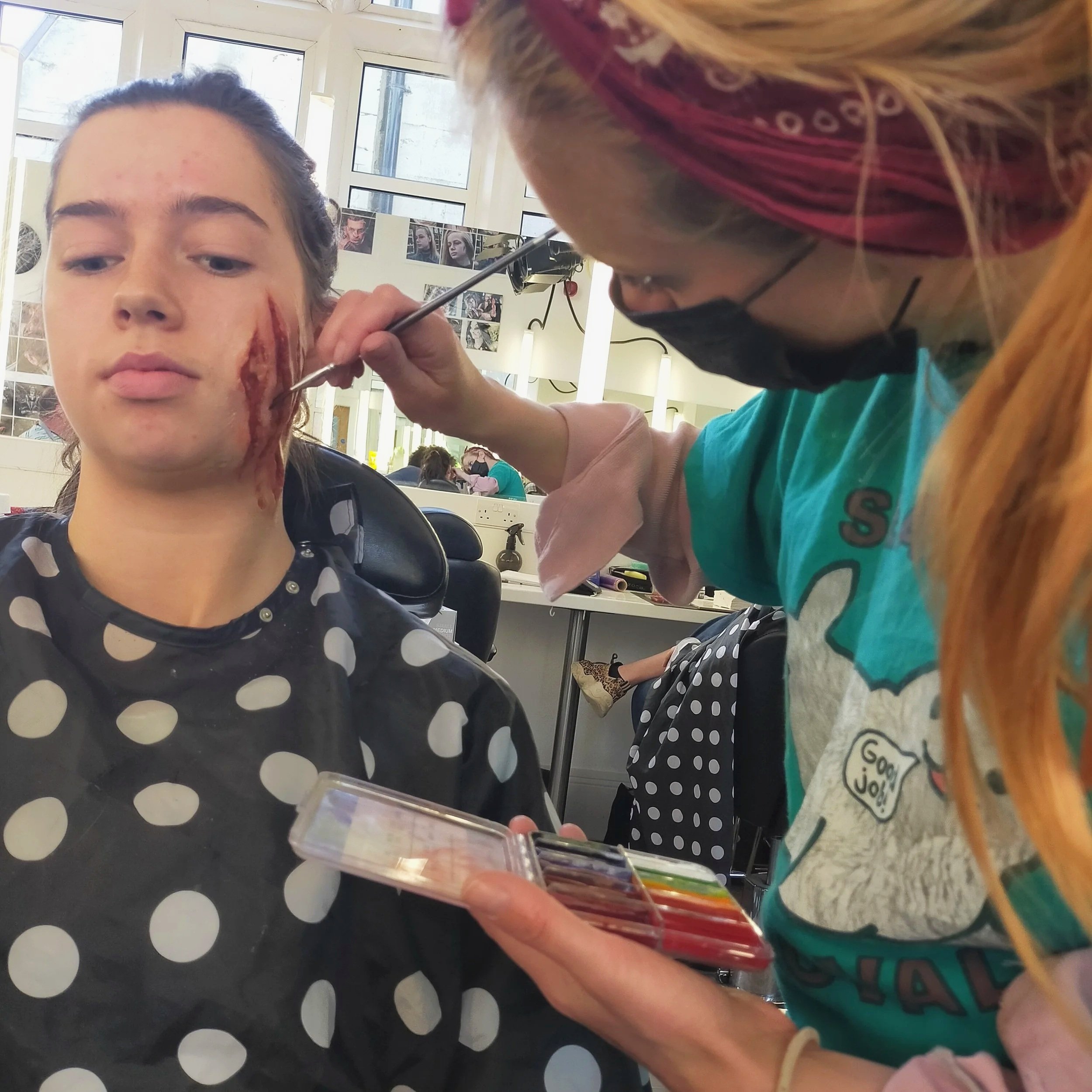 A person with blonde hair and a black face mask applies special effects makeup to a woman with dark hair and a black polka dot shirt, creating a bloody wound on her face in a salon or makeup studio.