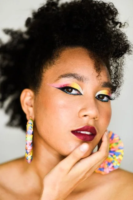 Close-up of woman with colorful makeup, curly hair, and rainbow earrings, touching her face.