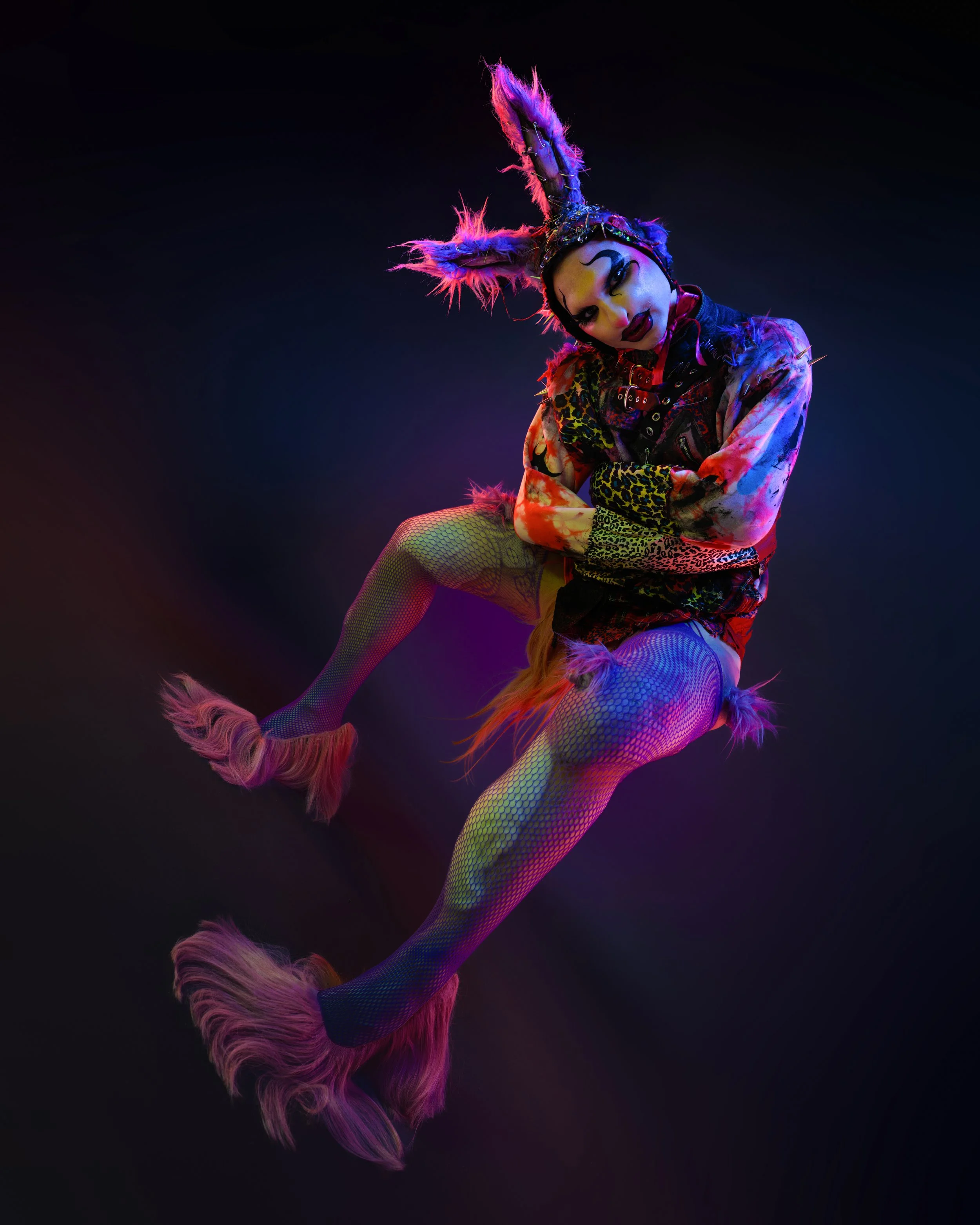 Person dressed in colorful, fantasy-inspired costume with rabbit ears, fishnet stockings, and furry boots, against a dark background with neon lighting.