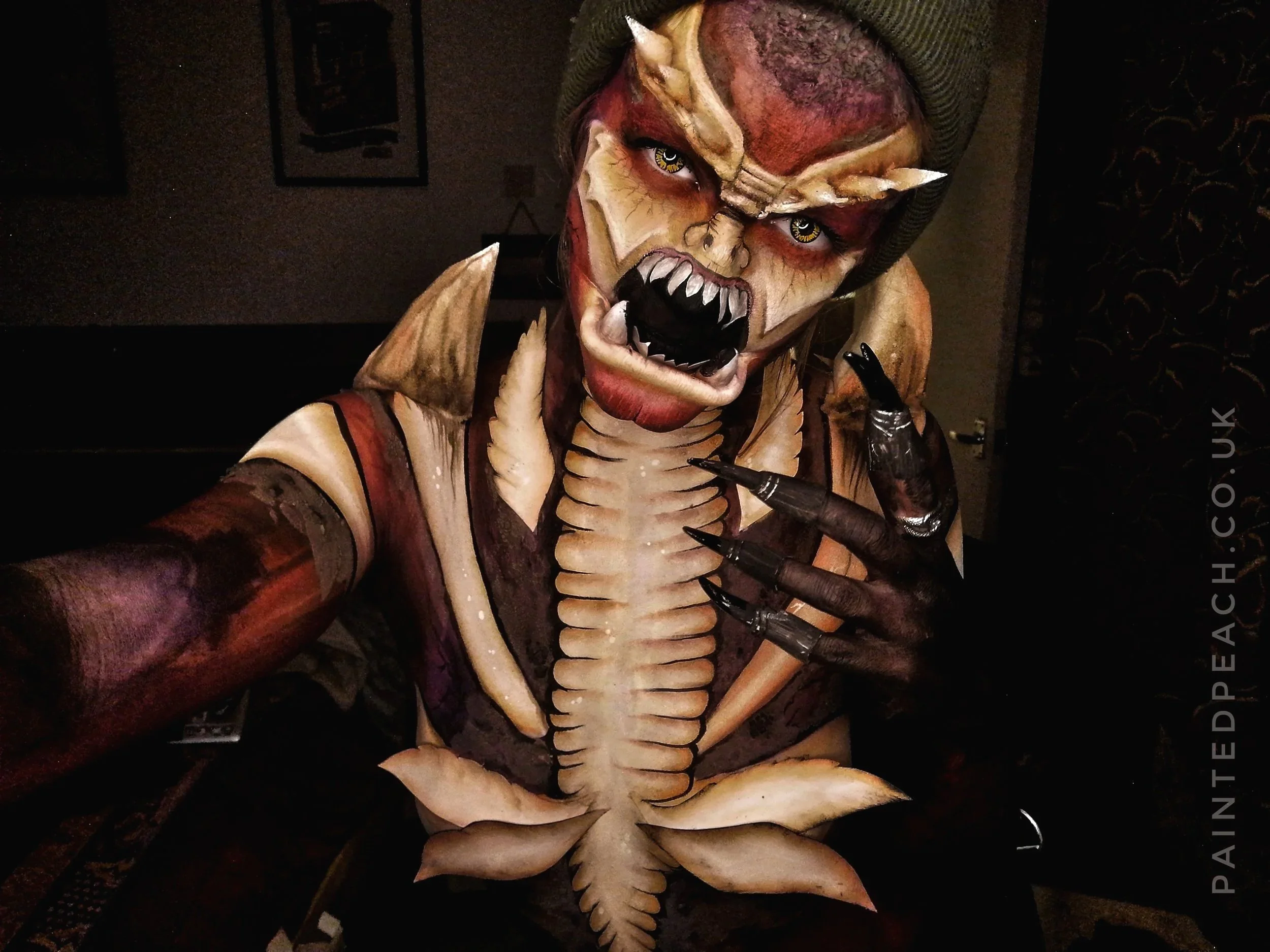 A person dressed in a horrifying, monster-like costume with detailed makeup and body paint, featuring sharp teeth, claws, and an intense facial expression.
