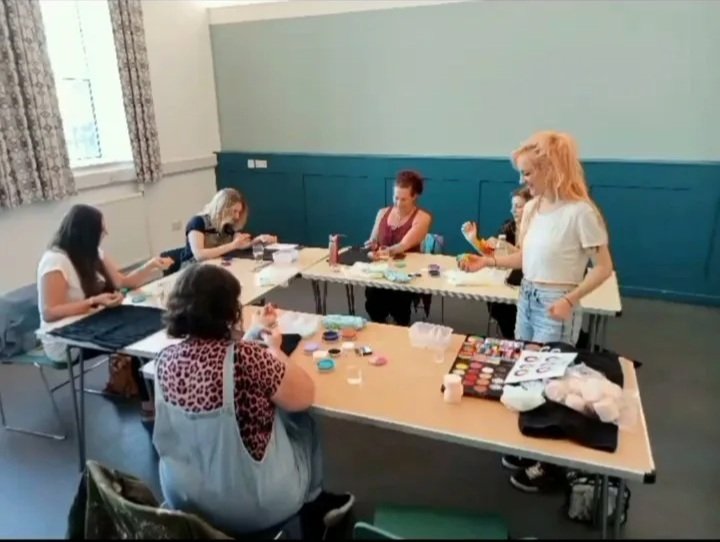 Group of women sitting at tables involved in a crafting activity, with art supplies and makeup palettes on the table.