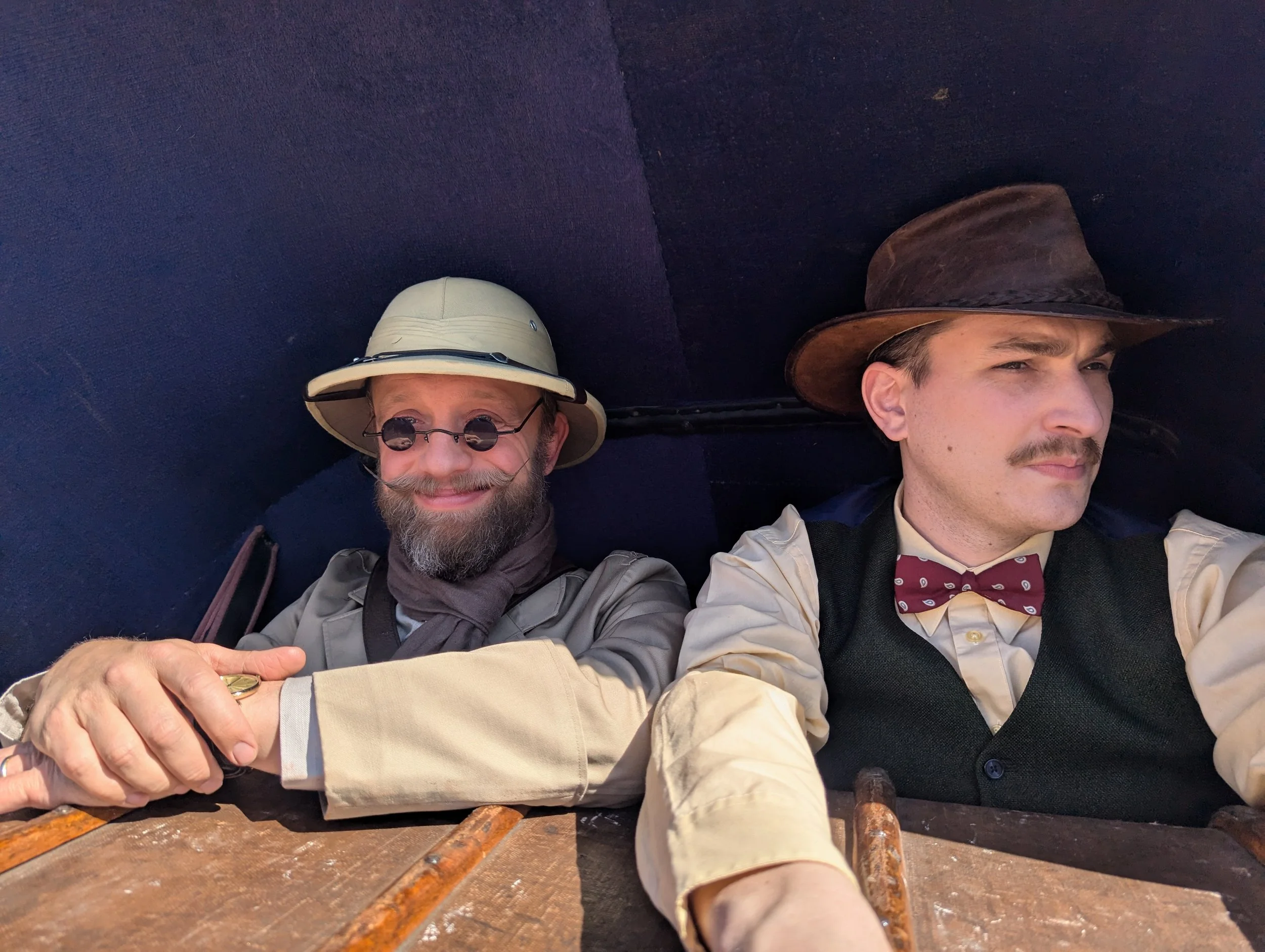 Two men dressed in vintage explorer attire, wearing hats and bow ties, lying on a wooden surface with a dark blue backdrop. One man is smiling and shaking hands, while the other looks serious and is looking away.