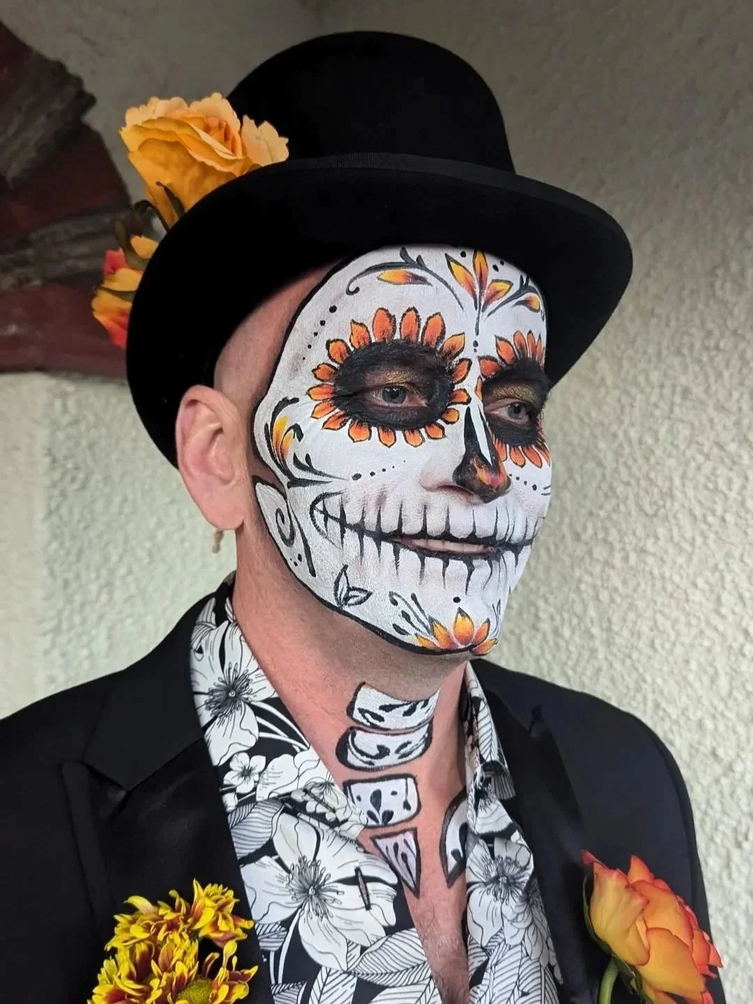 Person with sugar skull makeup, wearing a black hat with orange flowers, black jacket, and floral shirt, against a beige wall