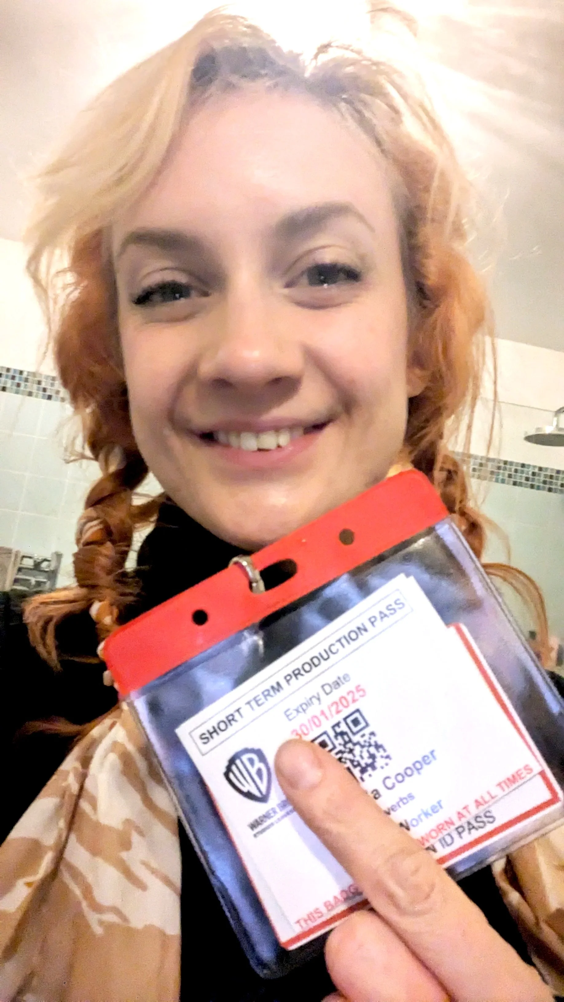 A person with curly, blonde hair holding a short-term production pass badge with a smile.