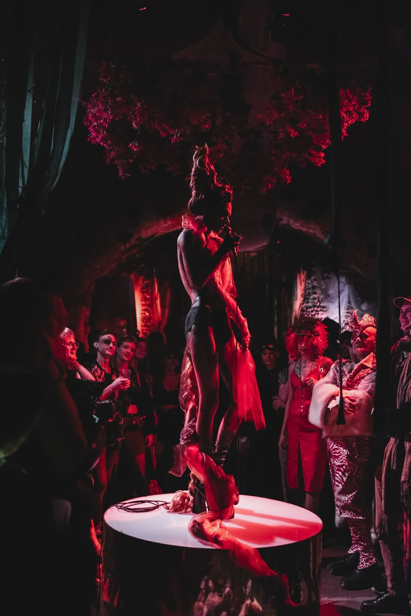 A performer stands on a platform with a microphone, surrounded by an audience in a dark, gothic-style setting. The scene is illuminated in red lighting, with theatrical decorations and performers in costumes, creating a dramatic atmosphere.